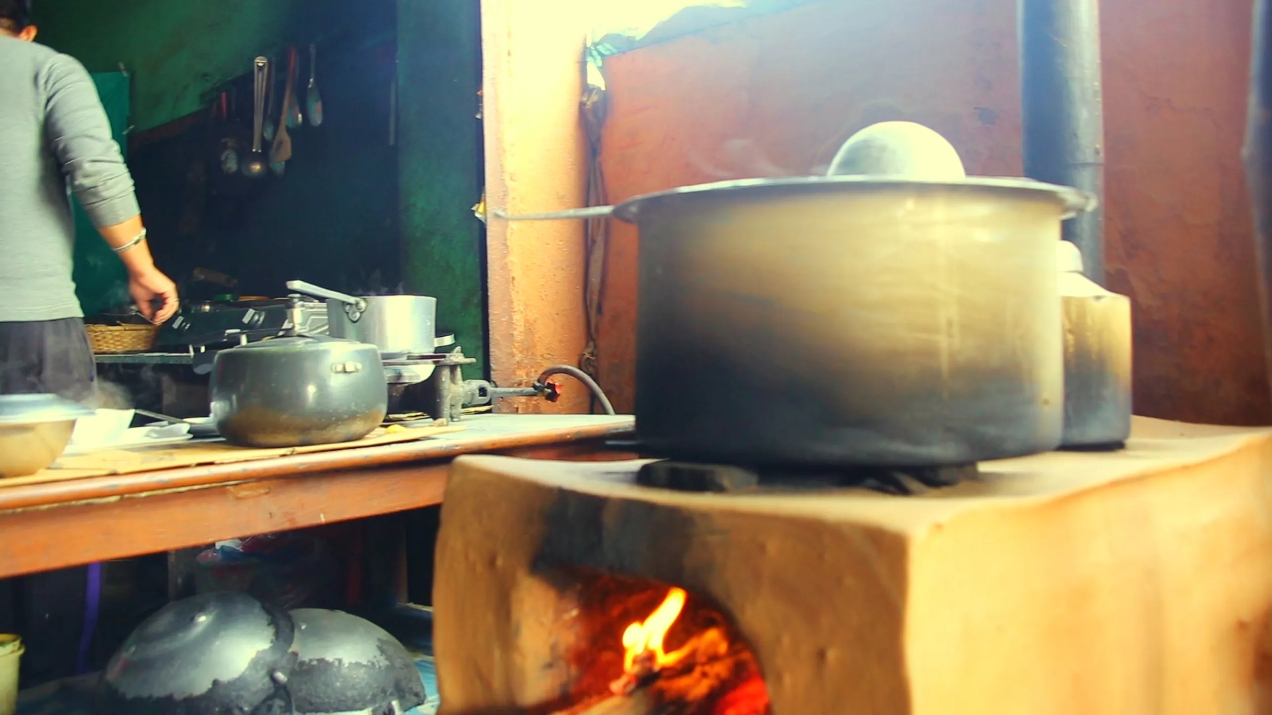 A close, atmospheric shot of a traditional wood-fire cooking stove in a rustic kitchen. The stove itself is a simple clay or brick platform, and within its open firebox, bright orange and yellow flames burn actively, the fire well-stoked and hot. Resting directly on top of the stove, a large, rounded metal pot or cooking vessel dominates the frame - darkened from long use, with condensation or steam forming at its lid and sides. The surface around the stove is wooden, worn, and darkened from years of smoke and heat. In the soft background, another stove or cooking area with additional pots is visible. The image is warmly lit from the fire itself, giving everything a golden, intimate glow. It is the quiet, ancient technology of mountain cooking.
