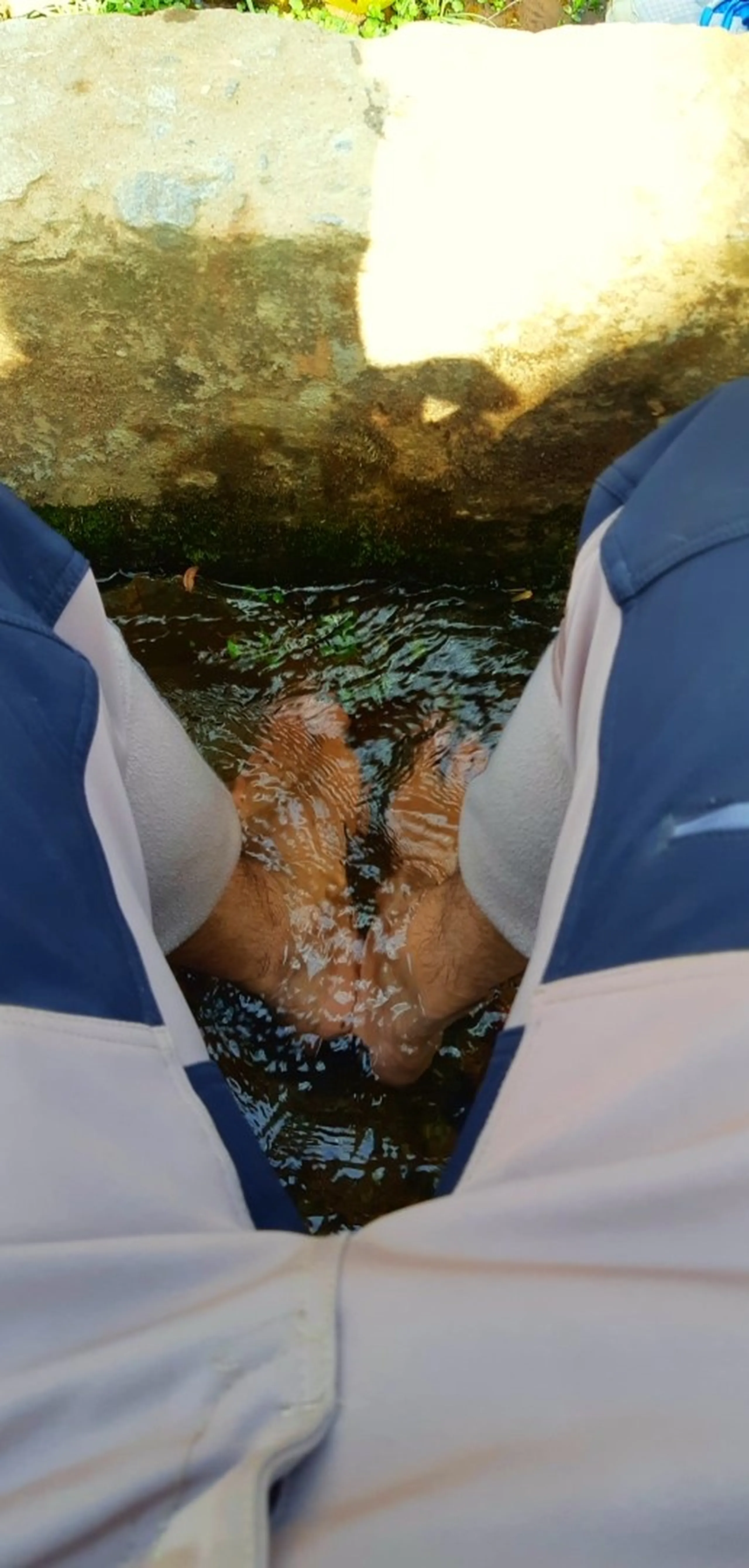 A first-person downward view of two bare feet submerged and splashing in a shallow, murky stream or irrigation channel. The water is dark brown and rippling around the feet. The person is wearing white pants (possibly school uniform trousers) with blue trim visible on the sides. The stream is bordered by rough stone or concrete edges, with green vegetation and bright sunlight visible in the background.
