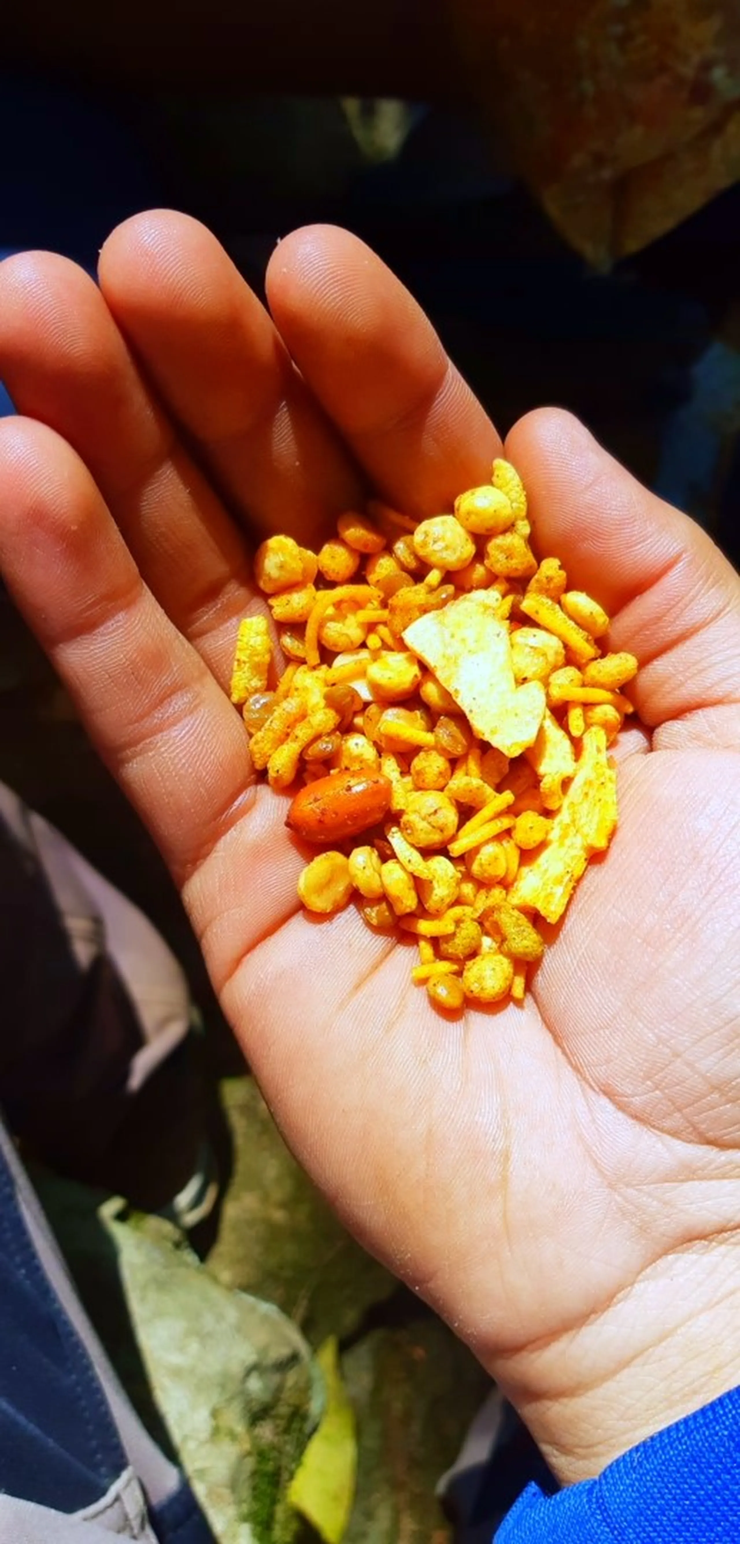 An open palm filled with a generous handful of small, bright yellow-orange puffed snack pieces - resembling spicy corn puffs or bhujia-style snacks - along with one larger chip fragment and a single reddish peanut. The snacks are vividly colored and densely packed in the center of the hand. The background is dark and out of focus, with a hint of blue sleeve visible.