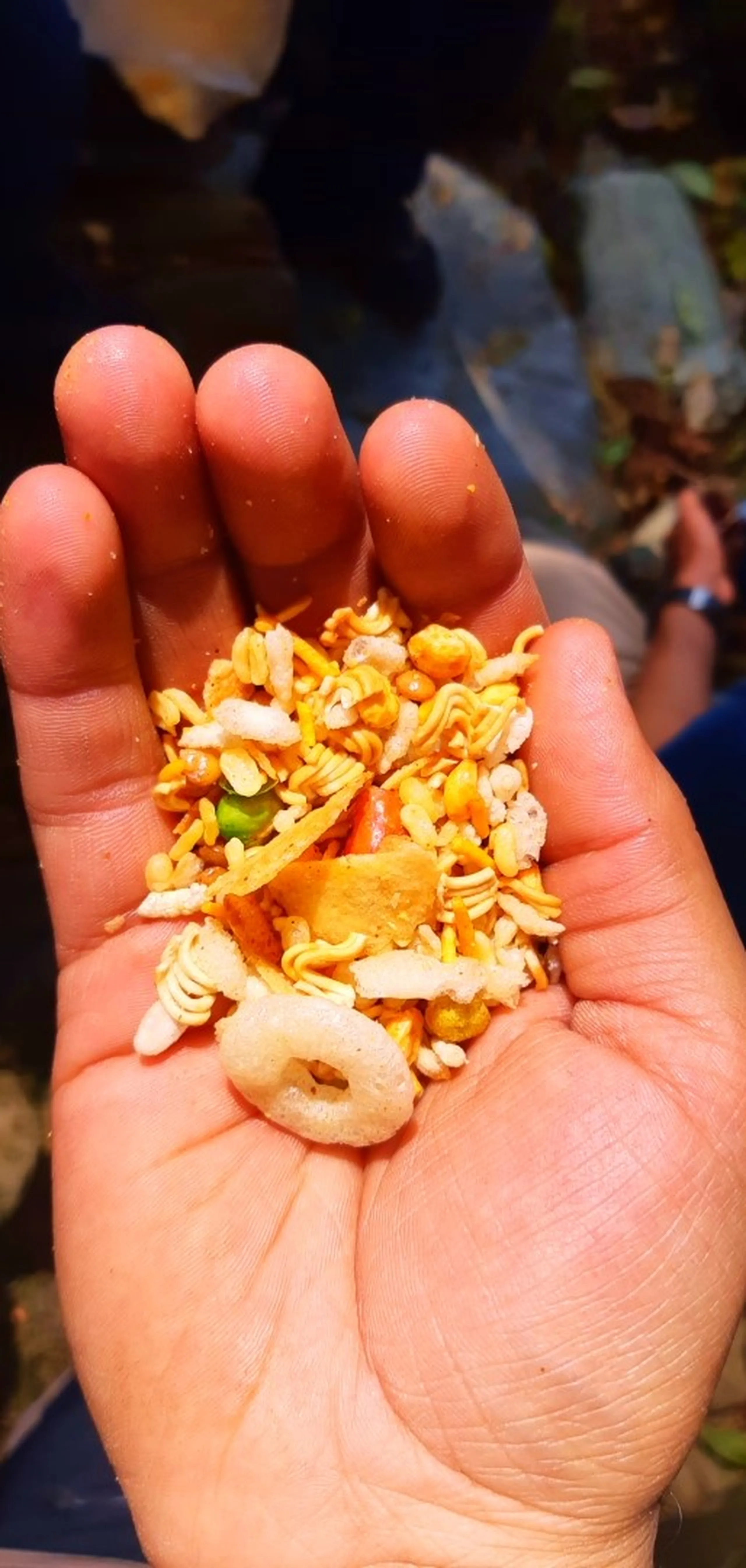A cupped hand holding a colorful mix of Indian-style snack mix (resembling chivda or Bombay mix), including yellow sev (thin fried noodles), small puffed pieces, a green chili, a round snack ring, and fragments of orange crackers. The hand is slightly damp or oily. The background is dark and blurred with hints of feet and a watch-wearing wrist of another person nearby.