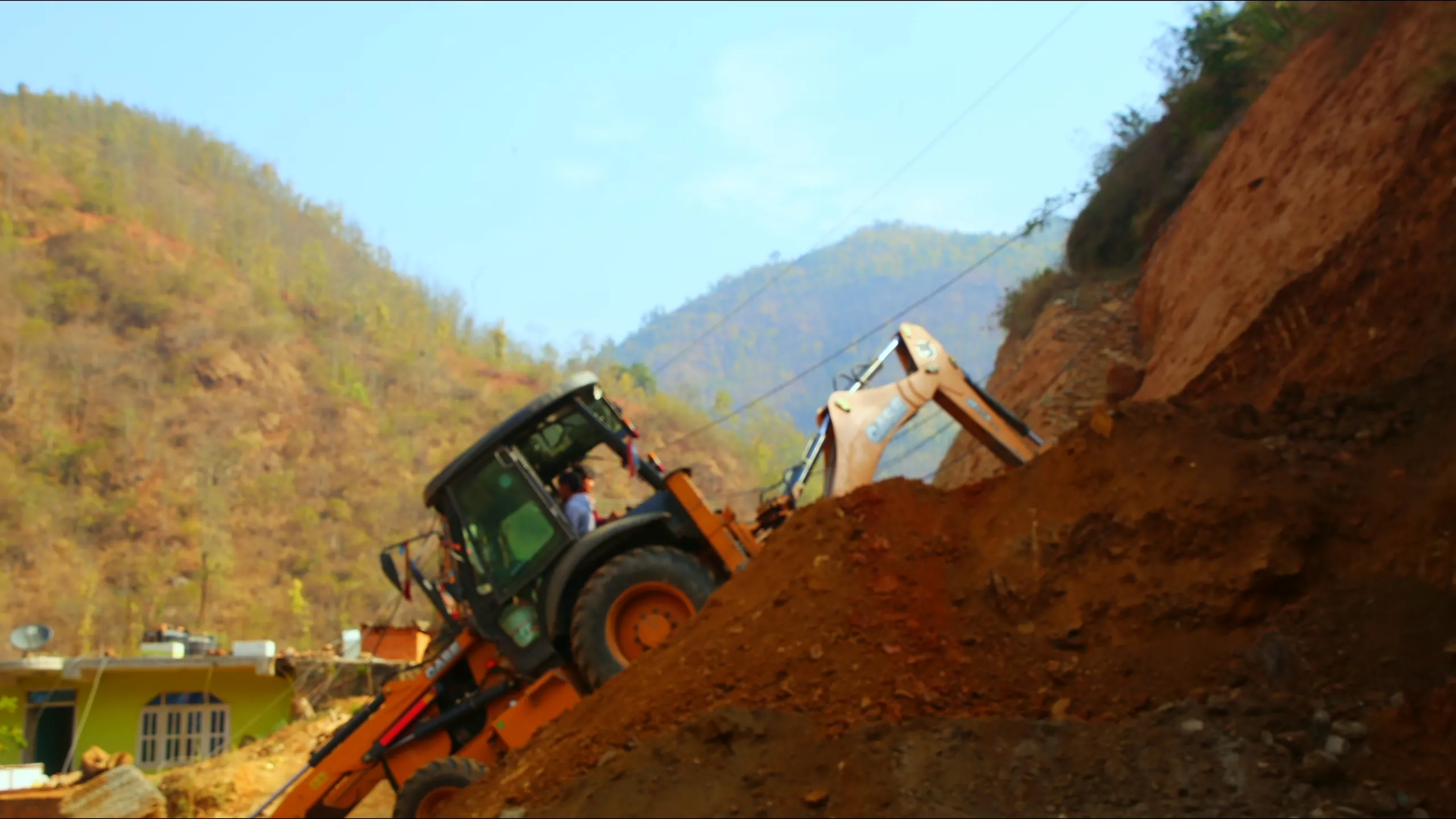 A wide landscape shot showing a green bulldozer or earthmover precariously positioned on a steep, red-clay hillside, mid-operation on what appears to be a road construction or landslide clearance site. A white vehicle is partially visible behind it. The hill in the background is covered in sparse dry trees, and the sky is a clear, bright blue. The scene has a rugged, remote mountain feel.