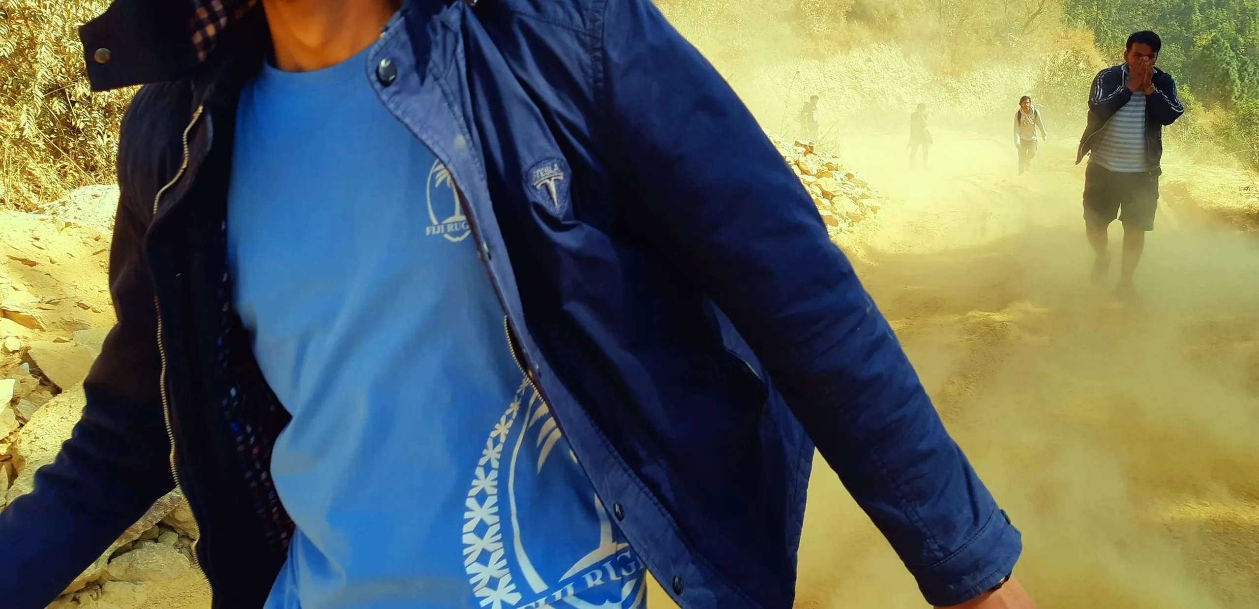 A motion-blurred action shot of a young man walking briskly toward the camera through a dusty, hazy environment. He wears a light blue graphic T-shirt and a navy blue jacket, and his expression is calm but purposeful. Behind him, another figure is visible in the dust cloud. The surroundings suggest a dry, earthen hillside or construction zone, with golden dust particles filling the air.