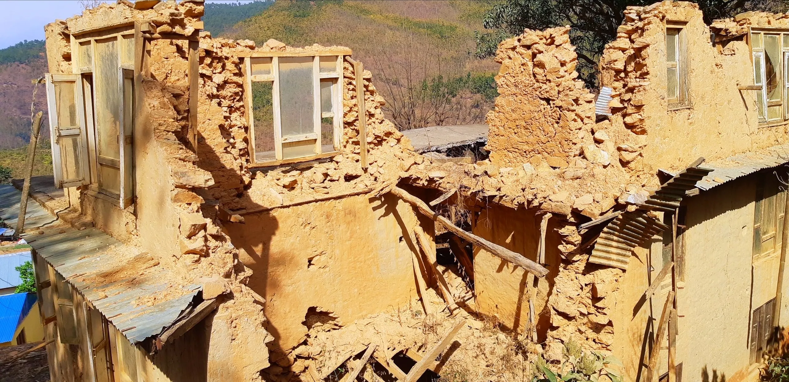 The ruins of a collapsed mud-brick or adobe building, with crumbling walls still partially standing. The structure shows exposed brickwork and broken window frames. The ground is littered with rubble and debris. In the background, terraced hillside fields and a calm blue river or lake are visible under a clear sky, suggesting a rural mountain village setting - possibly after an earthquake or natural disaster.