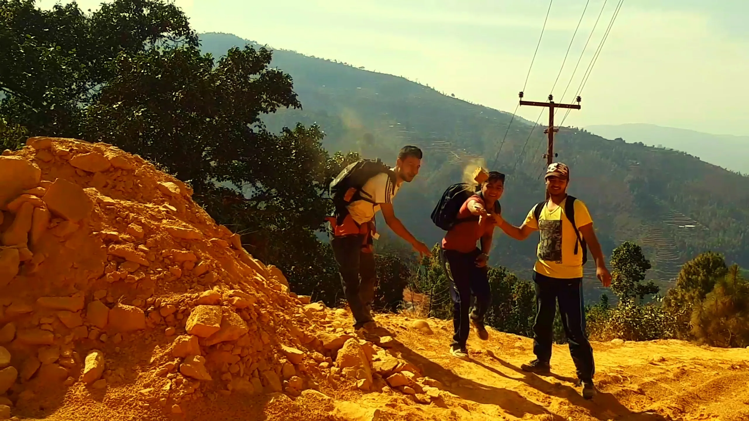 A group of three people hiking up a steep, reddish-orange dirt path or excavated hillside. They are carrying backpacks and appear to be mid-trek, with one person reaching back to help another. The terrain is raw and eroded, with bright orange-red soil. Electric poles and wires stretch across the background, and distant green hills are visible under a sunny, hazy sky.