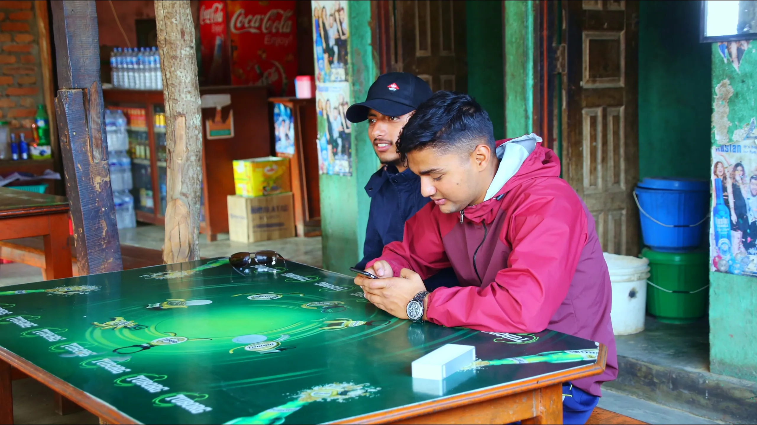 Two young men are bent intently over a green-topped game table outside a small roadside shop or teahouse. The table has a green surface printed with a football/soccer field diagram - this is a tabletop football strategy or board game. Both men are focused and engaged, studying the board carefully. One wears a red jacket, the other a dark cap and jacket. Behind them, the interior of the simple shop is partially visible - shelves with products, and a Coca-Cola branded sign or poster on the wall, a common sight in rural Nepali shops. The setting is informal and leisurely - a lazy afternoon game at the local shop. A white eraser or small object is visible on the table's edge.