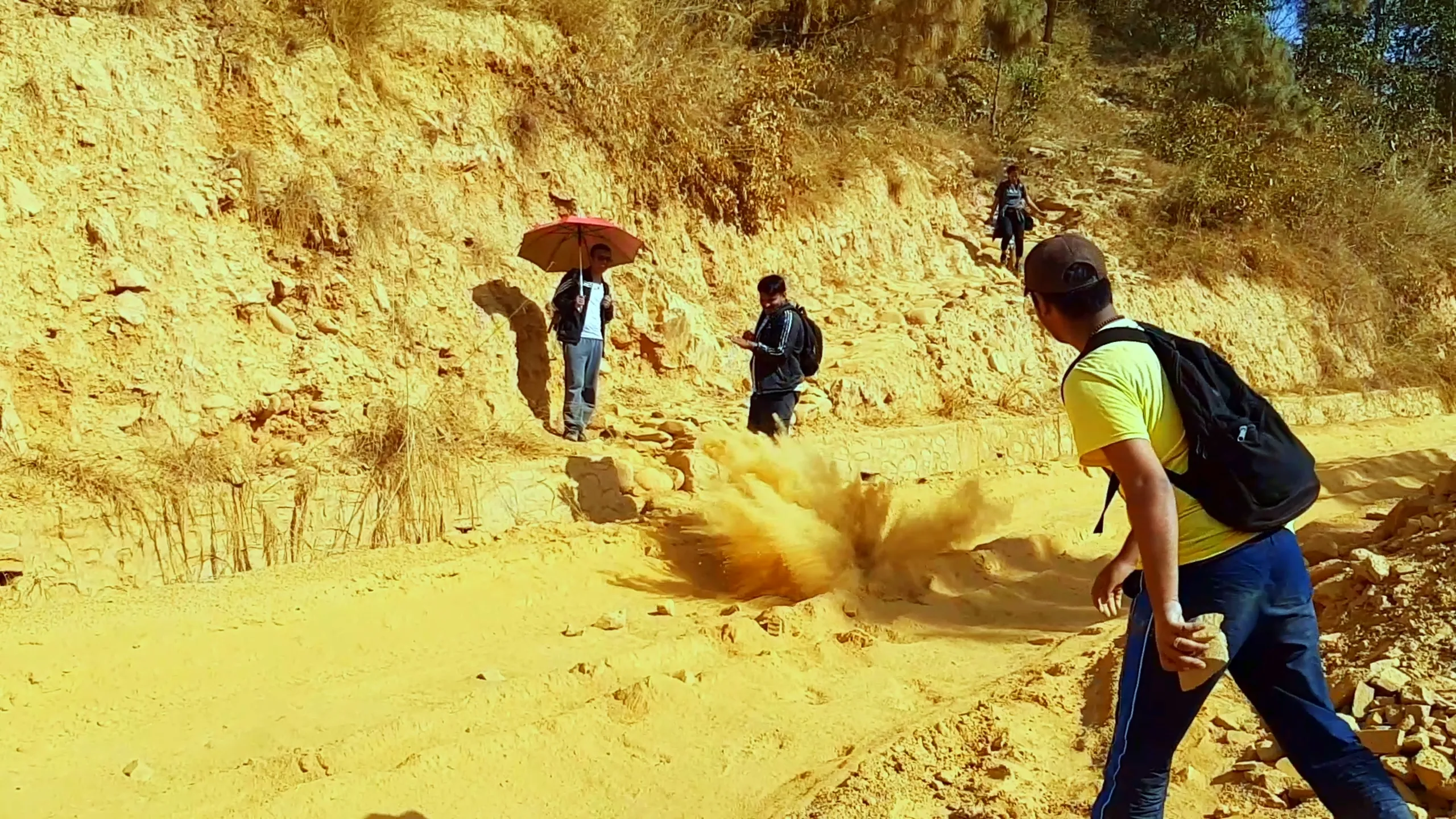 A dramatic outdoor scene on a reddish-yellow dirt hillside where a large cloud of orange-red dust has exploded from the ground, possibly from a landslide or deliberate ground disturbance. Several people are visible - one holding a red umbrella, others walking or standing nearby - appearing caught off guard. The dust plume fills the lower center of the frame in a burst.