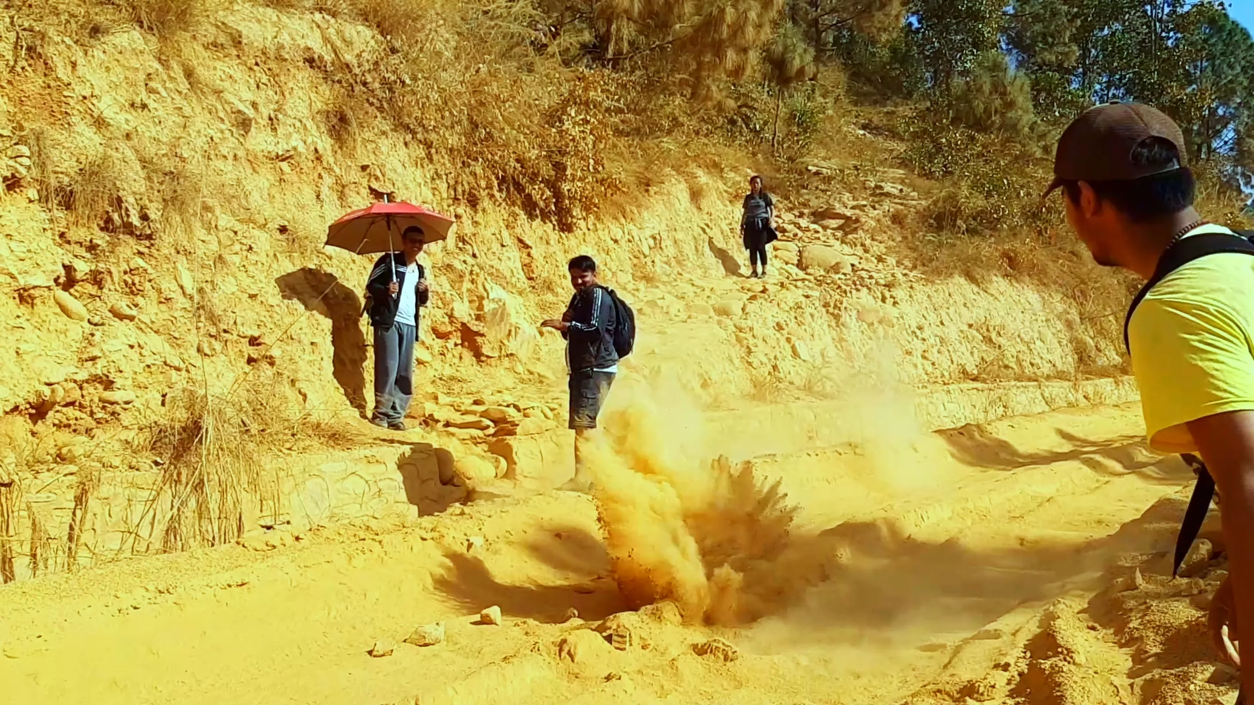 A continuation of the dust explosion scene: the orange dust cloud is now billowing larger and more dramatically. The person with the red umbrella is further back on the slope, and others are beginning to react and move. The red-clay hillside walls tower in the background, giving the scene a dramatic, almost cinematic quality.