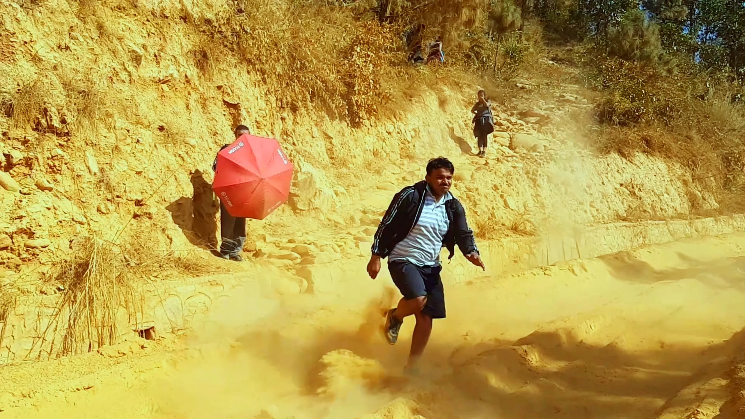 The dust cloud scene escalates - a man in a blue and white striped shirt and dark shorts is now running urgently toward the camera, away from the expanding dust burst behind him. The person with the red umbrella is visible in the upper background, and the red-clay cliffs frame the scene. The image captures pure motion and urgency.