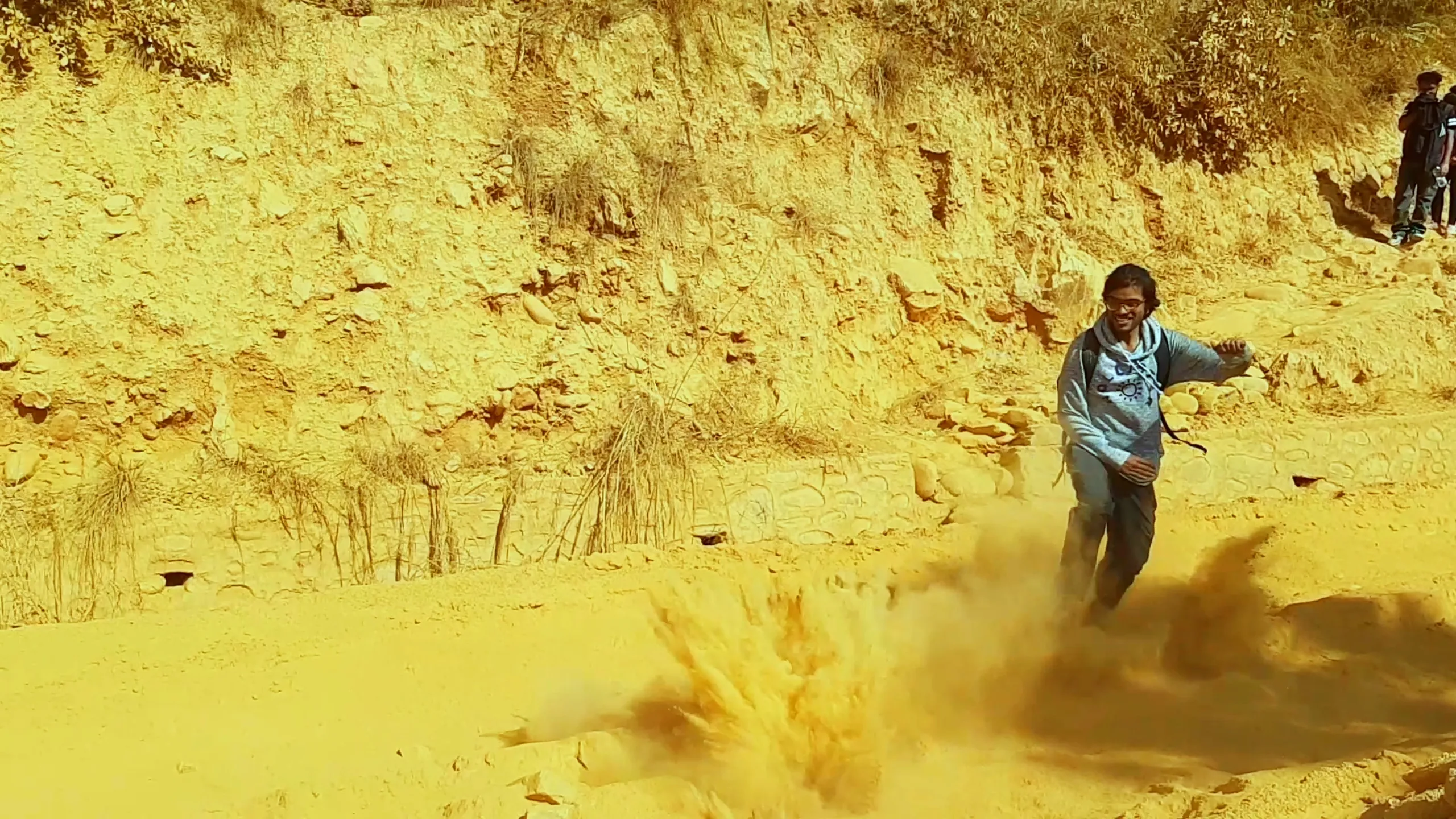 Peak action frame: a person in a light blue shirt is mid-sprint, running directly toward the camera with an expression of urgency or alarm. A fresh, violent burst of orange dust erupts from the ground right behind their feet, as if the ground is collapsing or exploding beneath them. The reddish earthen cliff walls surround the scene, amplifying the chaos.
