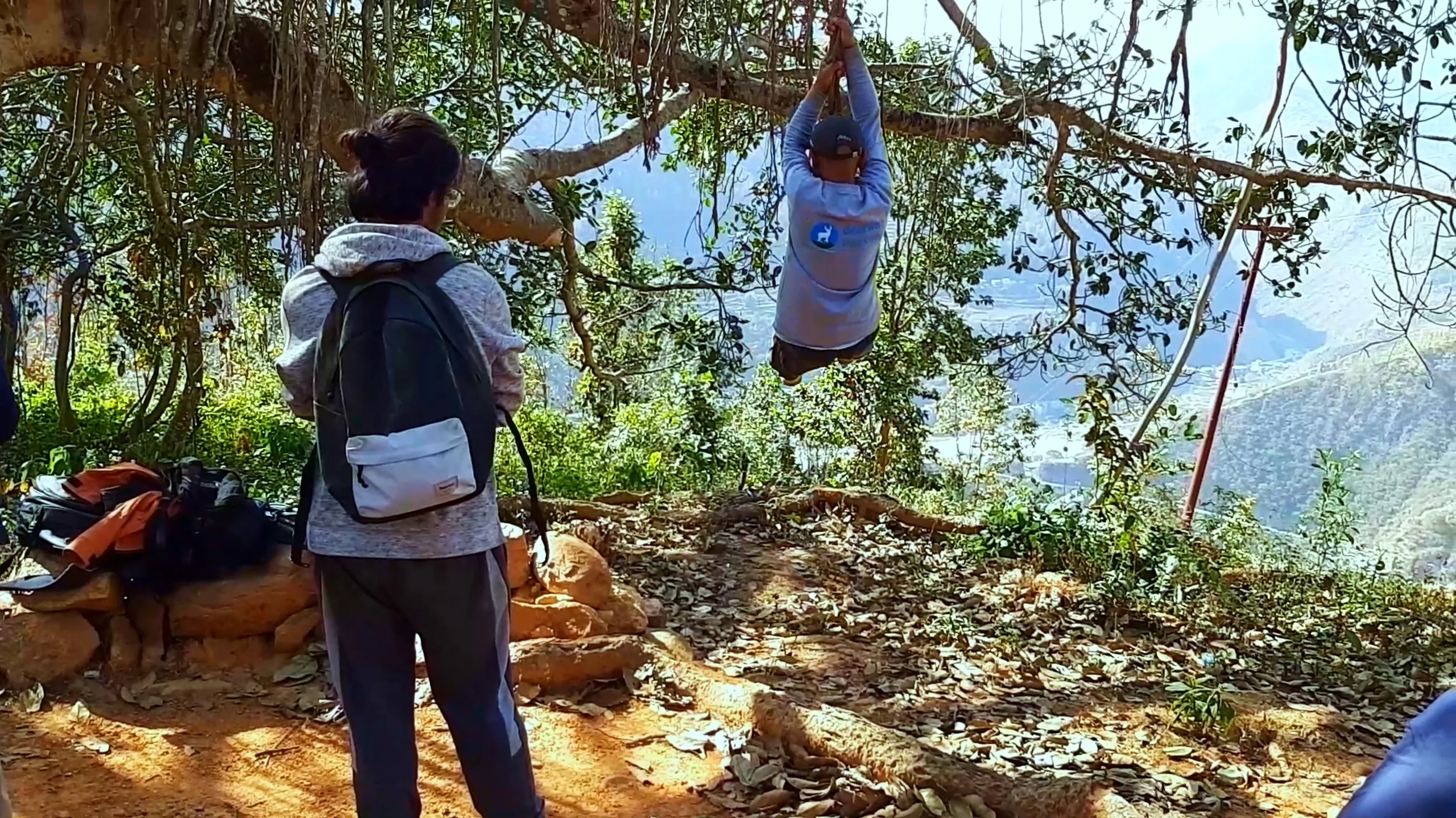 A person is swinging on a rope tied to a branch of the banyan tree, suspended mid-air against a backdrop of open sky and distant hazy mountains. Another person with a backpack watches from the foreground, their back to the camera. A dog is visible resting on the ground nearby. The scene feels spontaneous and free-spirited, like a break during a trek.
