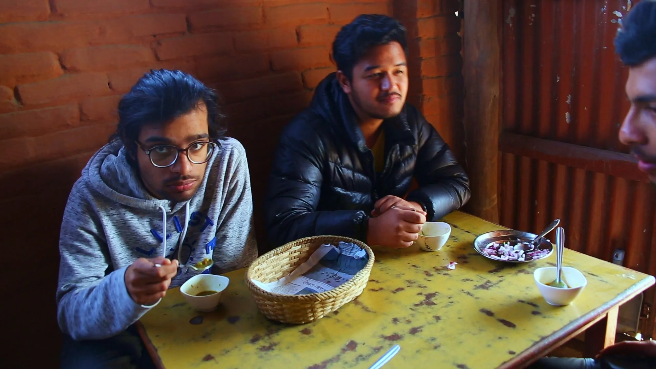Three young people sit together around a small yellow wooden table inside a simple, rustic room with dark red-brown brick walls. The left figure wears glasses and a light grey hoodie, holding a spoon and eating from a bowl - caught mid-meal, looking slightly toward the camera. The center figure wears a dark puffy jacket and looks forward with a neutral, relaxed expression. The right figure is partially visible, cut off at the frame edge. On the table between them, a small round wicker or cane basket sits in the center, and several bowls, small plates, and dishes with food are scattered around. The interior is dim and warm-toned. The mood is casual and companionable - friends sharing a simple meal in a local eatery.