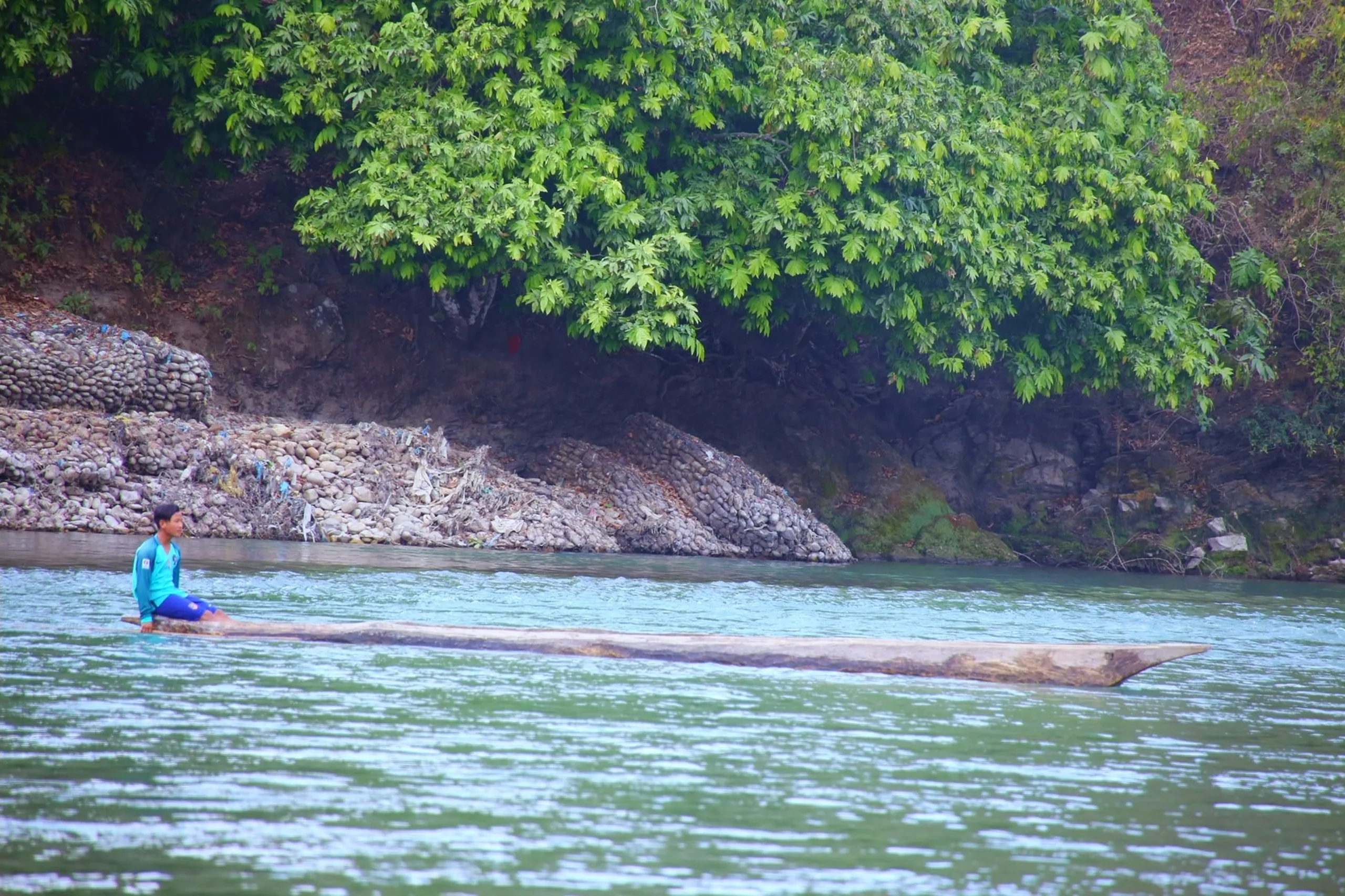 A lone figure dressed in a bright blue top sits or kneels on a long, flat wooden raft or log floating on a wide, calm turquoise-green river. The raft is low in the water and stretches horizontally across the frame. Lush green trees line the far bank, and large boulders are visible at the waterline. The river water is a beautiful clear teal color, and the scene feels remote, quiet, and timeless - like traditional river travel in a forested valley.