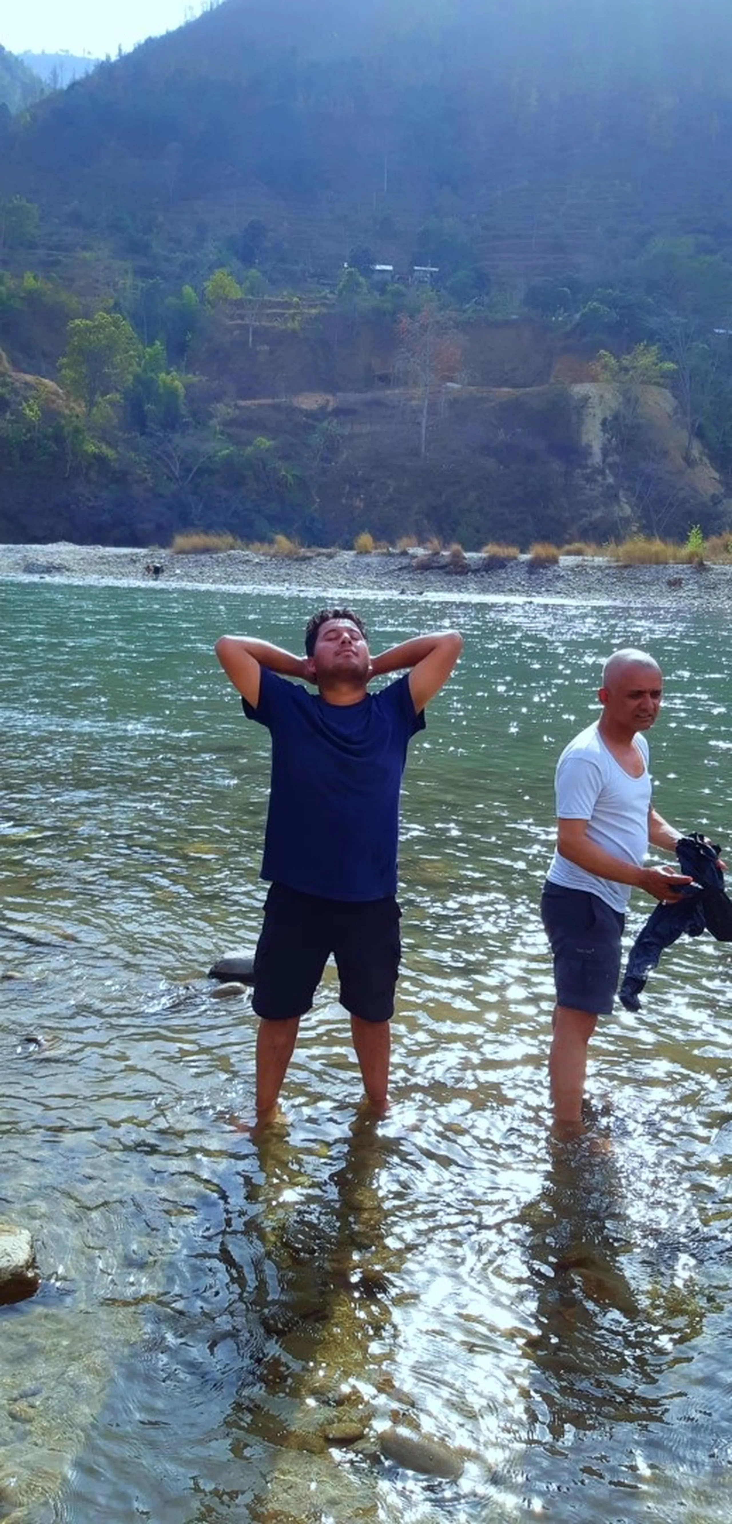 Two men are standing barefoot in the shallow, crystal-clear waters of a wide mountain river. The man on the left wears a navy blue T-shirt and black shorts, his head tilted back with eyes closed and hands clasped behind his head - the posture of someone experiencing total, blissful relief. The man on the right is bald, wearing a white vest, holding dark clothing in one hand. Behind them, a steep forested hillside rises sharply, with a glimpse of a small structure on the slope. The river water is transparent and gently rippling around their ankles.