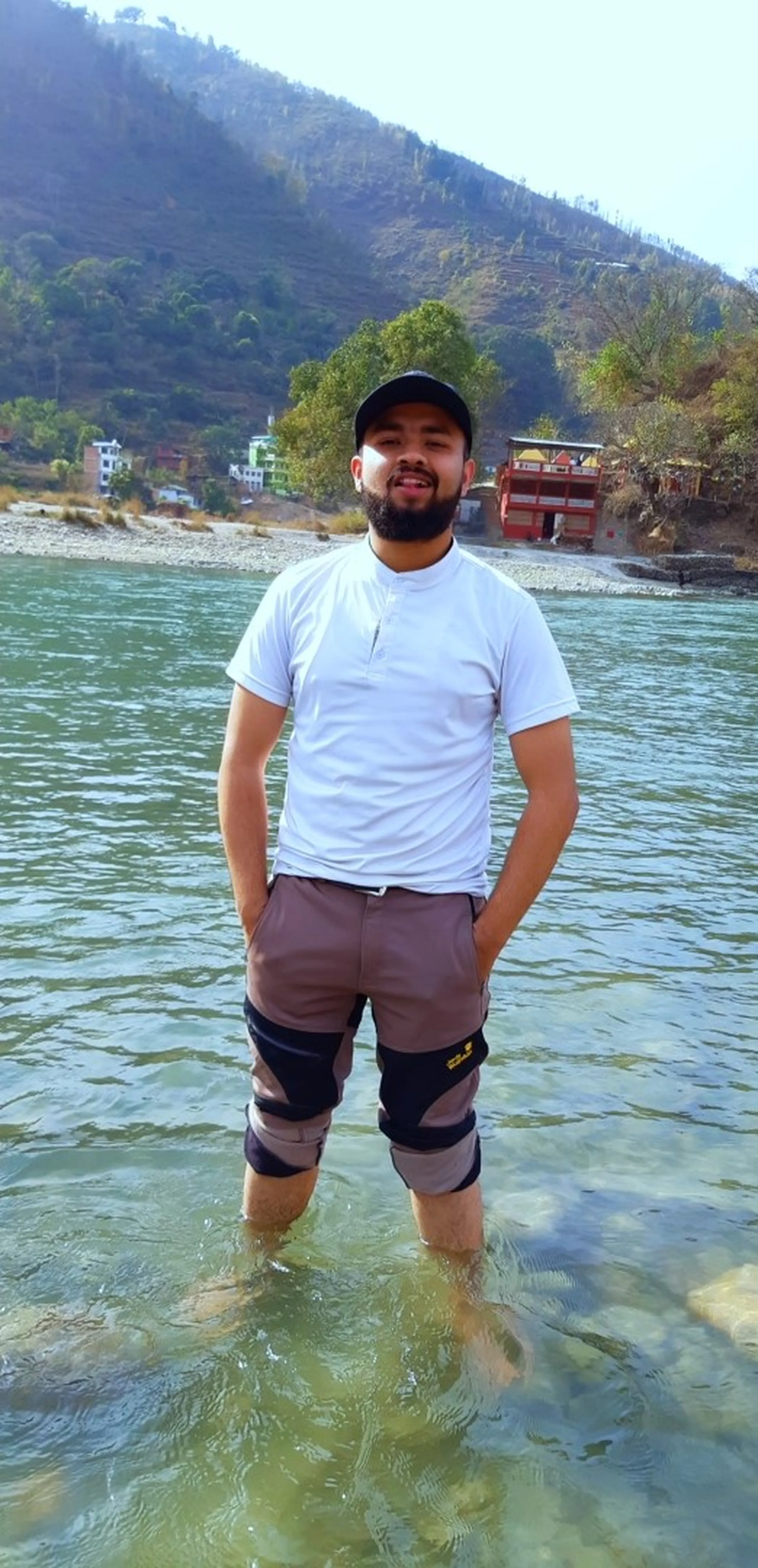 A young bearded man stands confidently mid-river, water reaching just above his knees. He wears a white T-shirt, a black baseball cap, and grey hiking trousers with black knee padding (branded with what appears to be Jack Wolfskin). His hands are in his pockets and he smiles warmly at the camera. Behind him, the wide river stretches across the frame, and a green forested hillside with a few colorful buildings - including one in red - lines the far bank. The scene is bright, sunny, and cheerful.