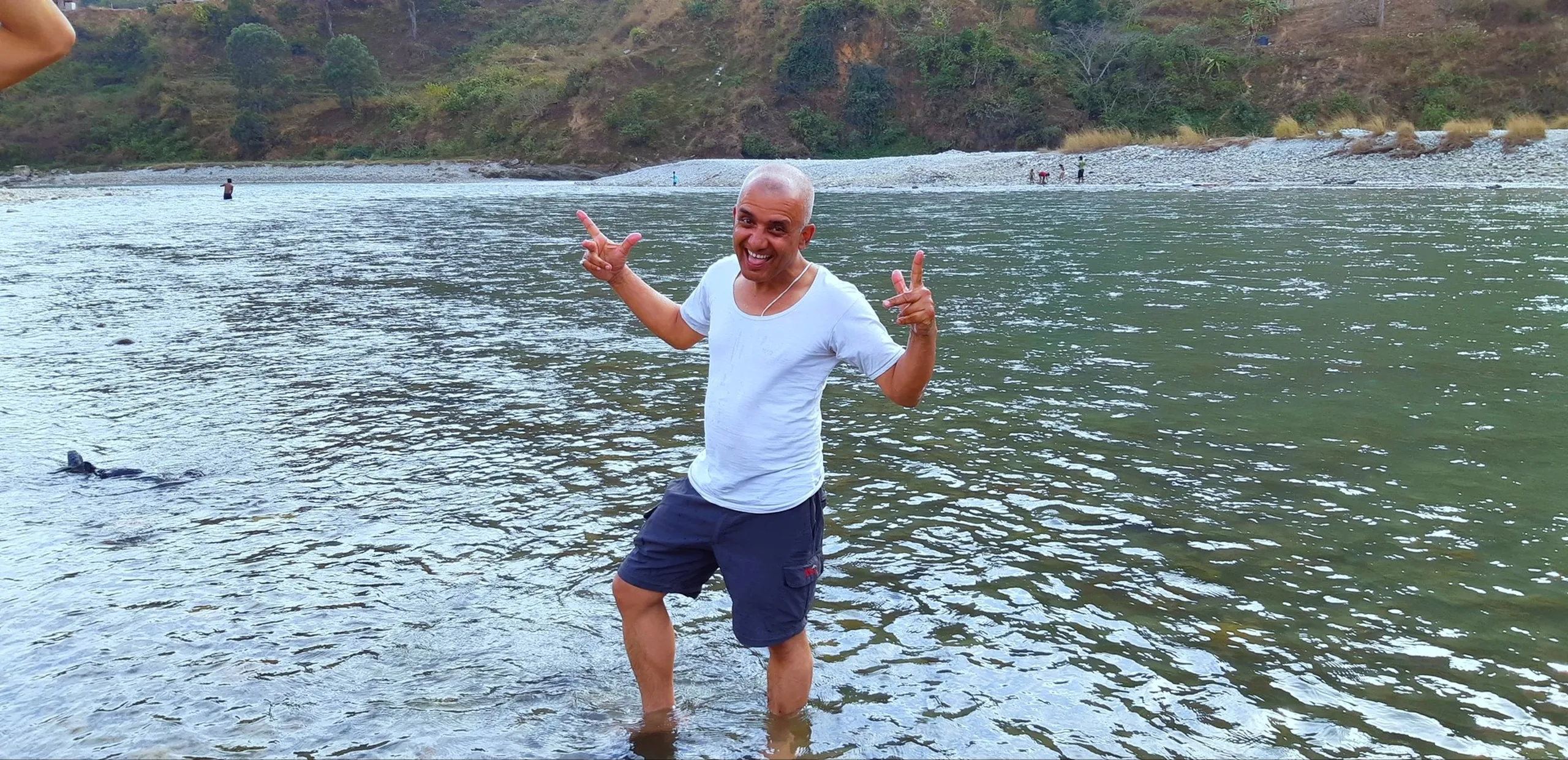 A bald man stands ankle-deep in a wide, shallow river, facing the camera with a huge grin and both hands making thumbs-up gestures. He wears a white T-shirt and blue shorts, visibly overjoyed. The river behind him is calm and wide, with a mountain hillside in the background, partially in evening shadow. The mood is pure, unrestrained happiness.