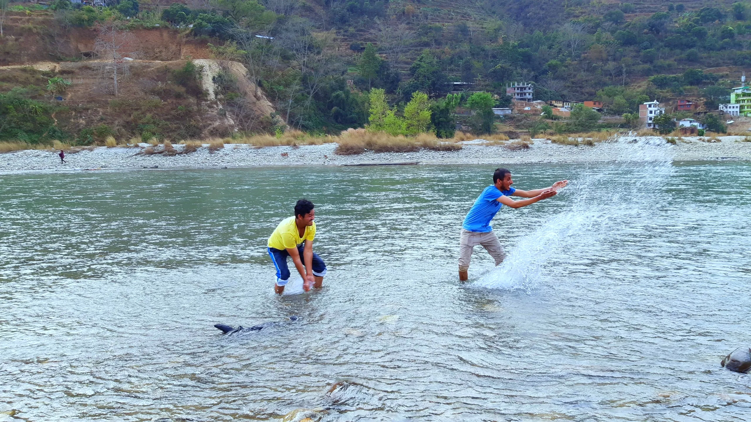 An action shot of two young men playing and splashing in a shallow river. One wears a yellow top, the other a blue shirt. One appears to have just slapped or kicked up a large splash of water toward the other, who is leaping or stumbling in reaction. The water sprays dramatically around them. The far bank shows eroded red-brown hillsides and a small cluster of buildings. The image is full of spontaneous fun and energy.