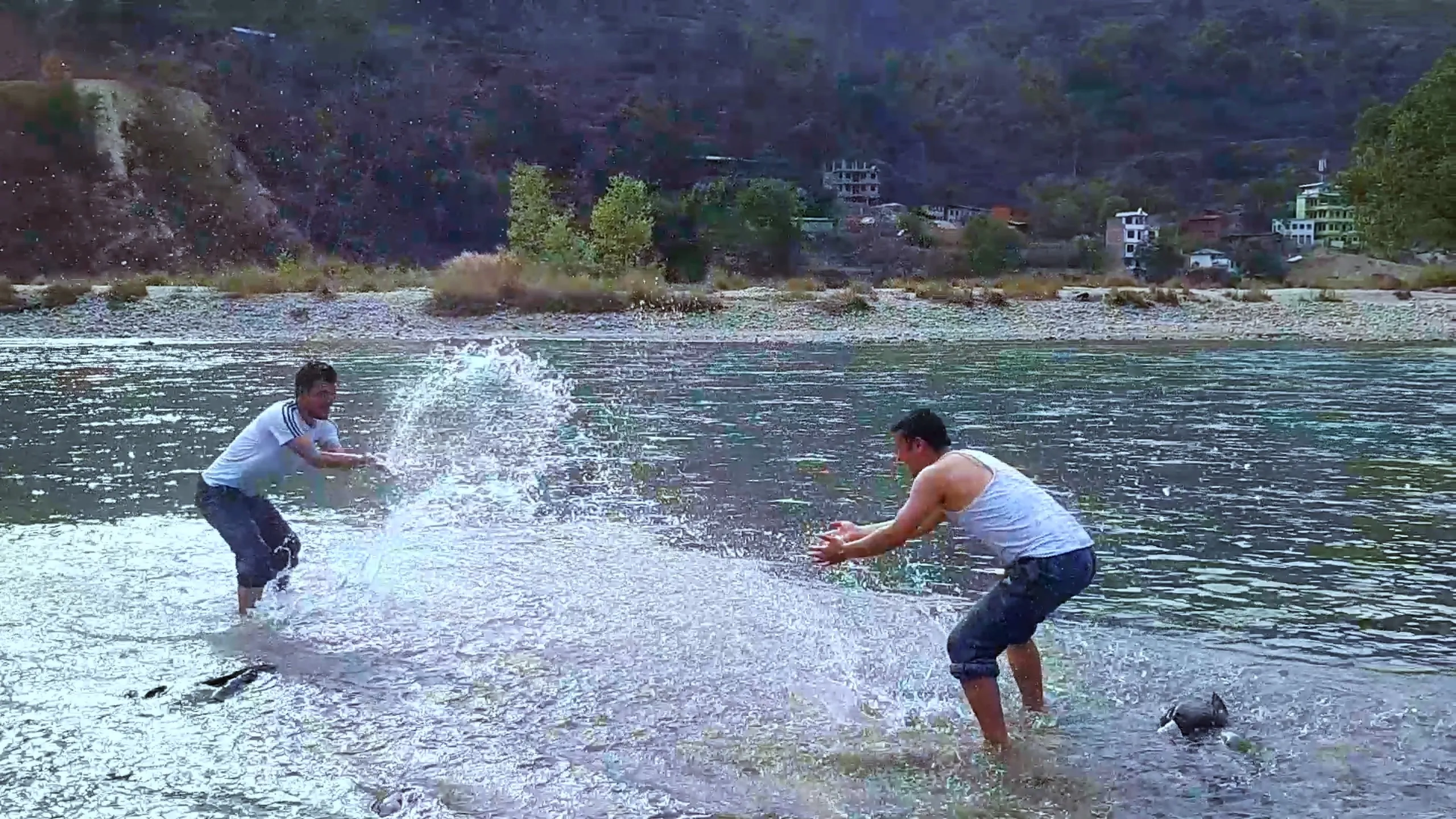 Two men are engaged in an all-out water fight in the shallows of the river. One in a white shirt and one in a white sleeveless vest are both bent forward, arms outstretched, flinging massive arcs of water at each other. A huge spray of white water fills the center of the frame. The background shows forested slopes and a small riverside settlement. The moment is chaotic, joyful, and high-energy.