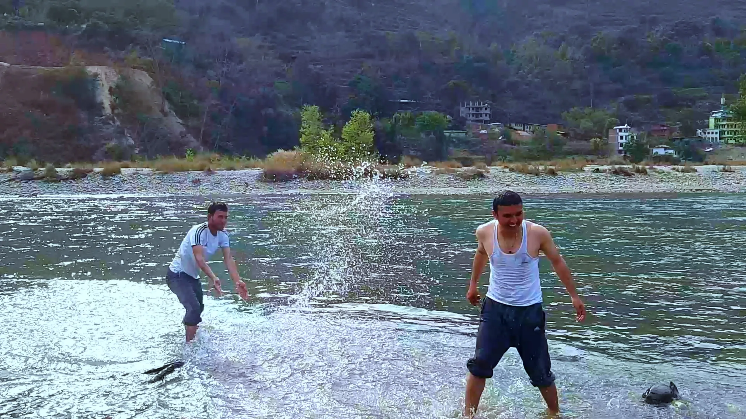 The water fight continues - the same two men are now caught in an even more dramatic splash. The man in the white vest is bent low, flinging water with both hands, while his companion leans back in response. Water is flying in all directions, creating a dramatic white burst between them. The hillside and riverside town are visible in the cool blue-grey background. The scene has a playful, almost battle-like intensity.