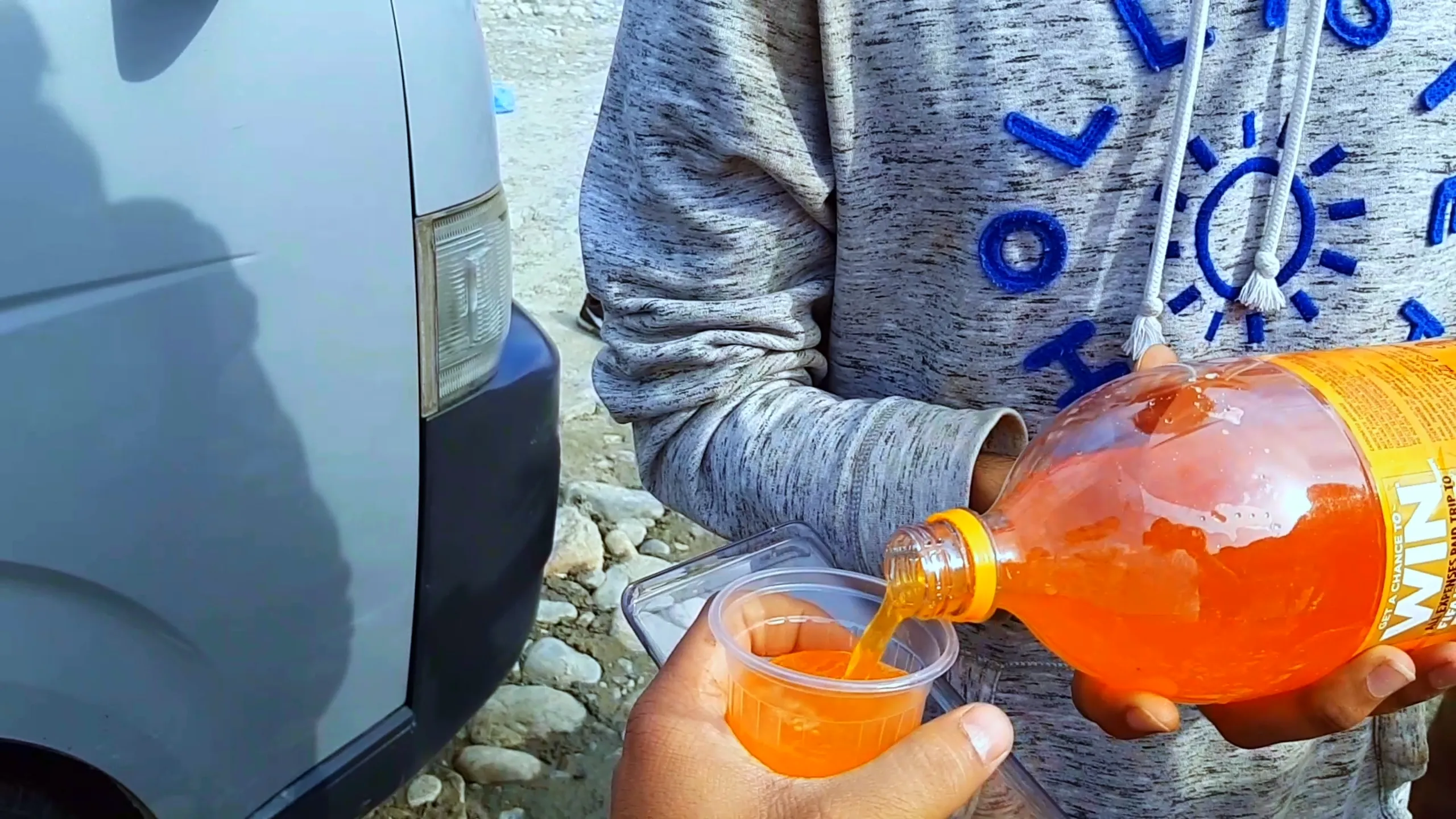 A close-up shot of someone pouring a bright orange fizzy drink from a large plastic bottle into a small clear plastic cup. The person wears a grey hoodie with blue text. The pour is mid-action, the orange liquid catching the light. The setting appears to be outdoors near a vehicle - a grey car door is partially visible. The ground appears gravelly. The image has the feel of a roadside refreshment break during a trip.