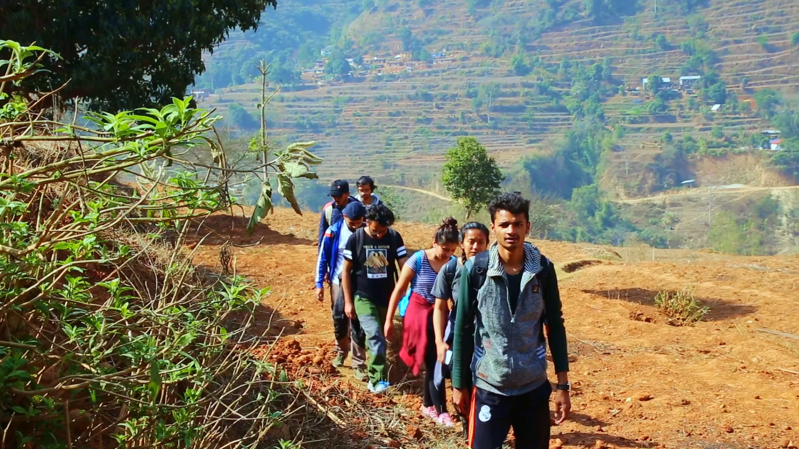 A line of six or seven trekkers moves single file along a narrow red-dirt trail cut across a sunny hillside. They are strung out one behind the other in a classic trekking formation, all moving in the same direction along the path. Their clothing is casual - a mix of everyday jackets, t-shirts, and light trekking gear in various colors including blue, black, and grey. The hillside around them is dry and red-earthed, with sparse low vegetation. In the background, rolling hills are terraced into agricultural steps, the fields golden-brown and dry. The landscape is warm and open, the sky bright. The image captures the simple, rhythmic pleasure of walking in a group through the hills.