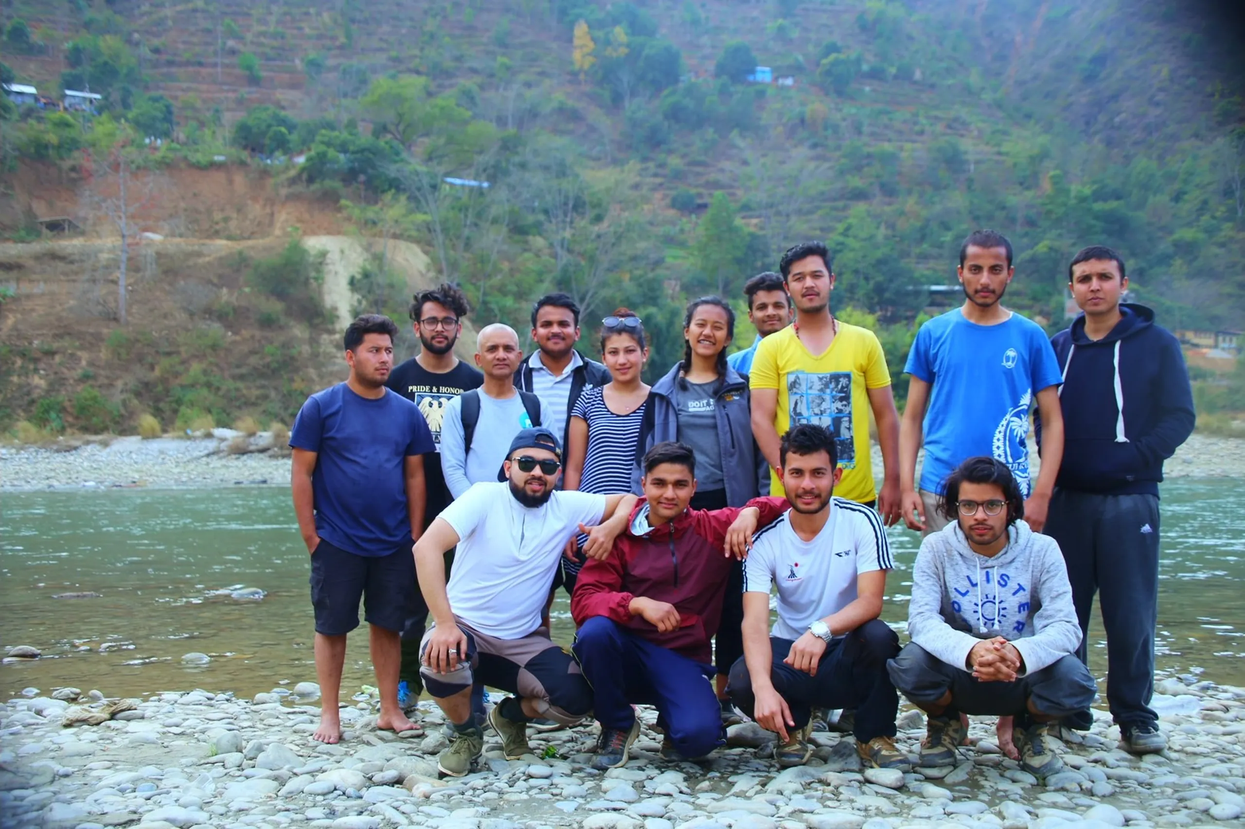 A group photo of approximately 13-14 people - men and one woman - posing together on the rocky bank of a mountain river. Some are standing, others crouching in the front row. They wear a variety of colorful casual clothes - blue, yellow, grey, green. Everyone is smiling. Behind them, the river flows past and terraced hillsides with sparse trees and a small settlement rise in the background. The atmosphere is warm and celebratory, like an end-of-trip group shot.