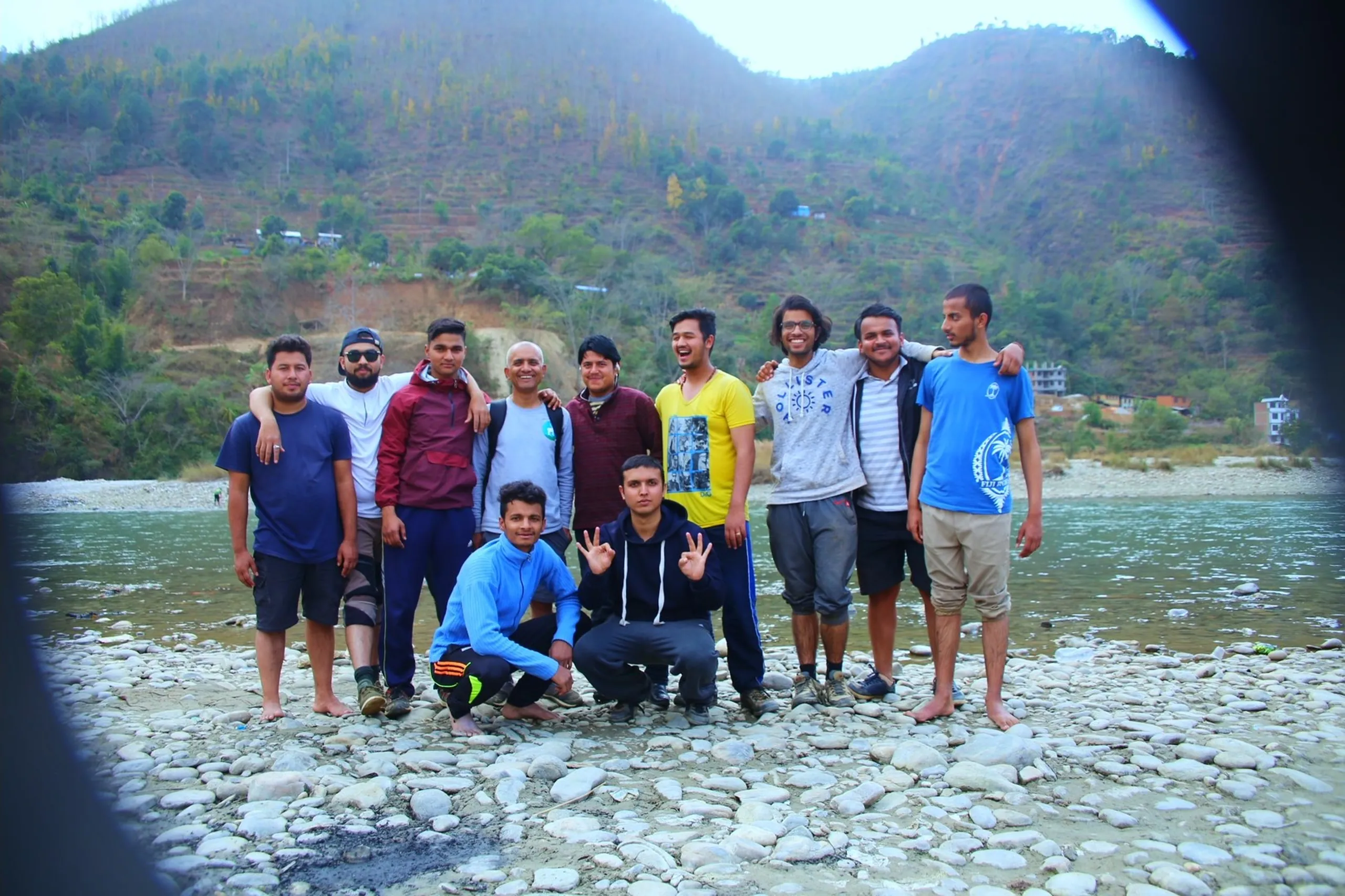 A second group photo of about 11 people on the same rocky riverbank, this time shot from a slightly different angle - possibly through a vehicle window, as a dark curved frame partially borders the left edge. Two people crouch in the front, others stand in a row behind. One person makes a peace sign. The forested hillside and the river are visible behind them. The mood is relaxed and happy.