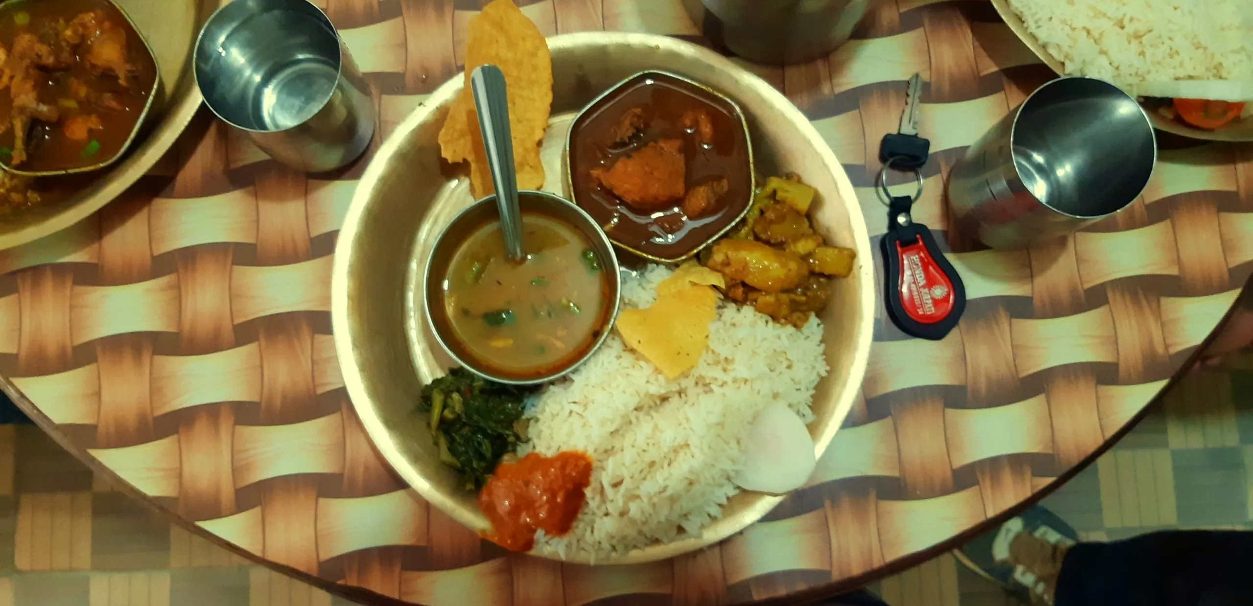 A top-down overhead shot of a traditional South Asian thali meal served on a large round white plate placed on a woven rattan or cane-patterned table surface. The plate contains a mound of steamed white rice in the center, surrounded by small portions of various curries and sides: a dark meat or lentil curry, a golden curry, a green vegetable dish, a small chutney or pickle in red, and a small steel bowl of dal soup. A spoon is resting in one of the dishes. A steel tumbler, a car key fob, and the edge of another dish are visible around the plate.