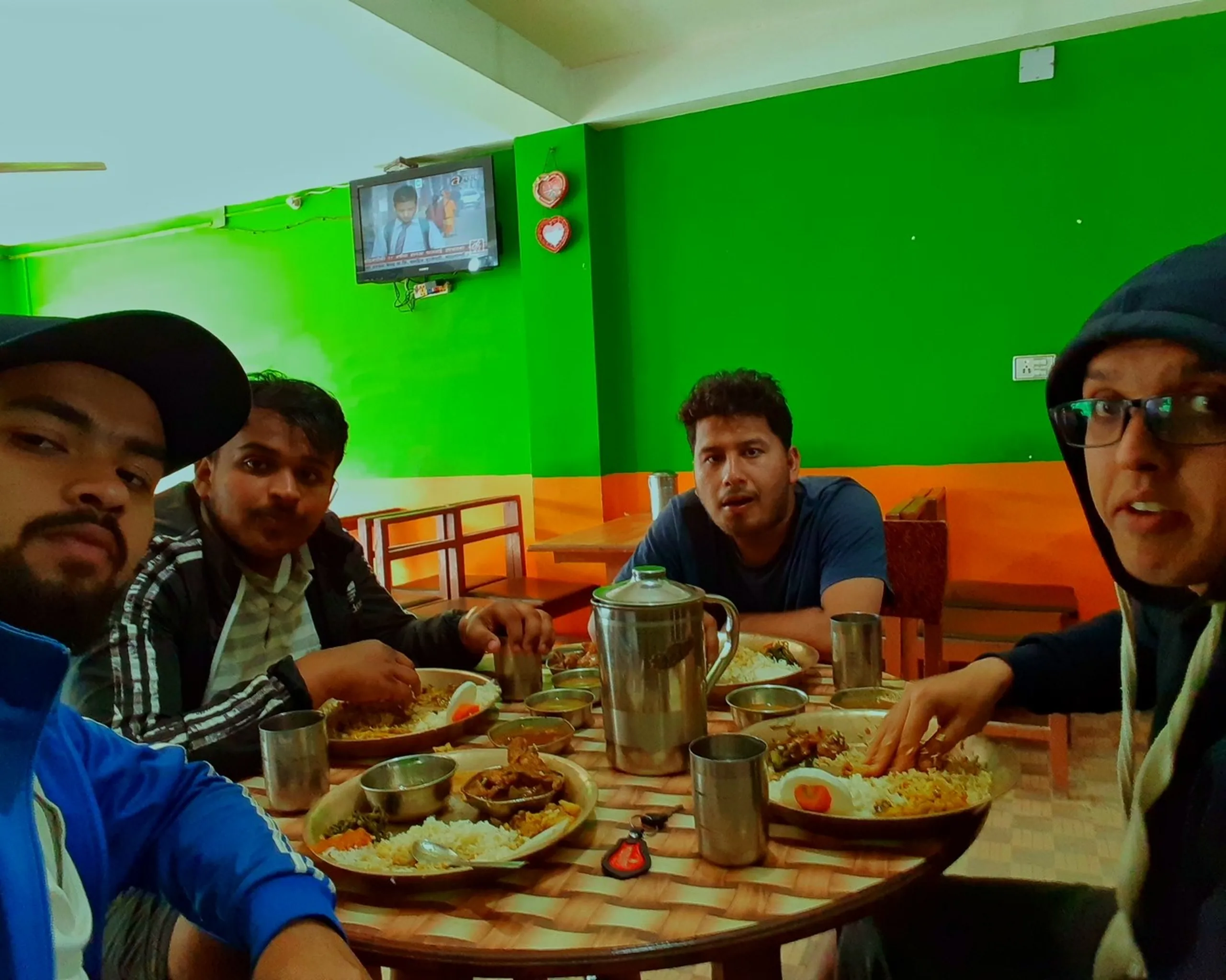 Four young men sit around a wooden dining table inside a small, casual restaurant or dhaba. The walls are painted in bold green and orange, and a small wall-mounted television is visible in the upper background. The table is crowded with steel plates of food, a steel jug (likely water), and cups. The men are mid-conversation - one is leaning forward speaking, others are listening or reacting with animated expressions. The scene feels like a lively mealtime on a road trip.