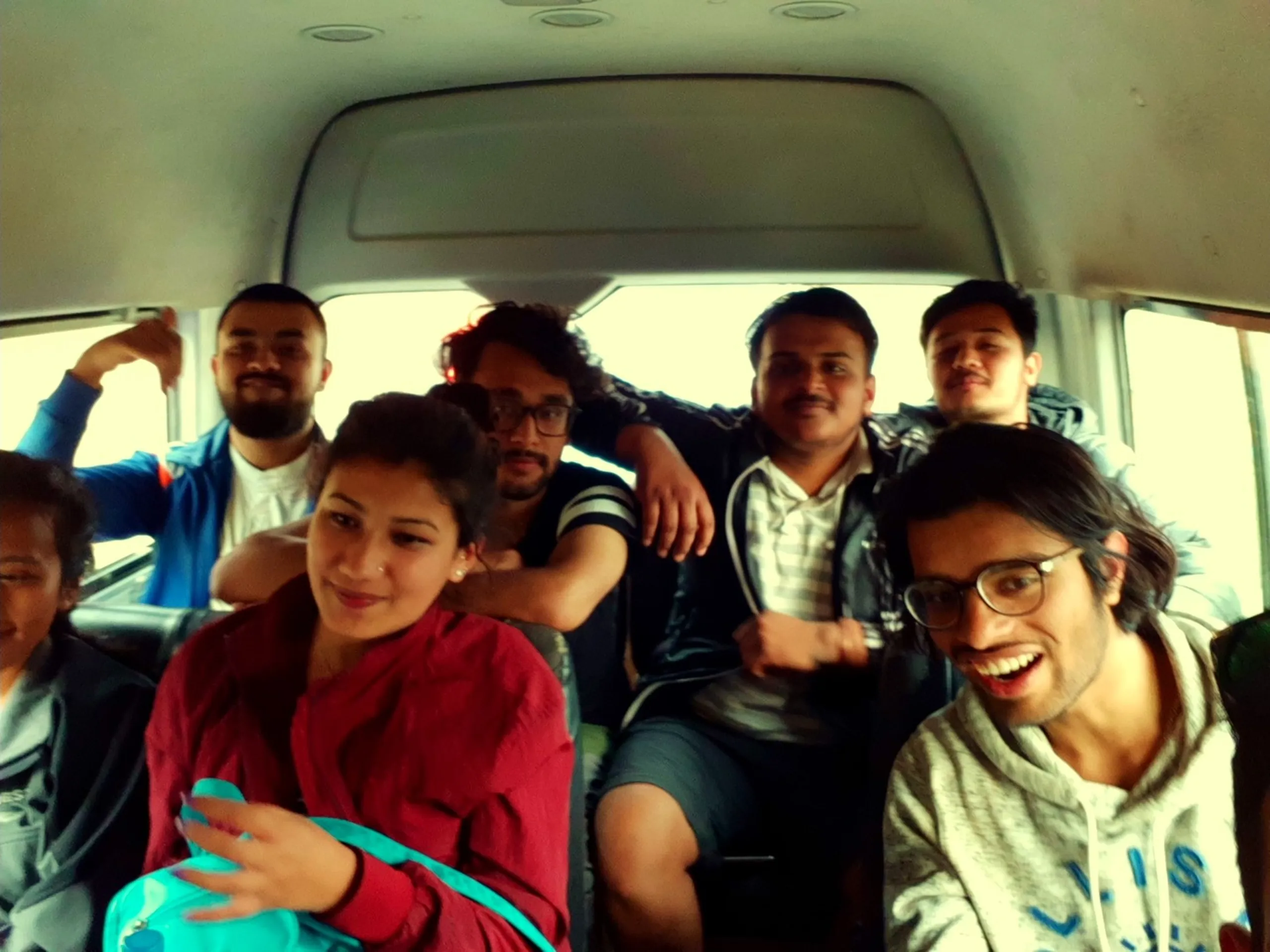 The interior of a minivan or microbus packed with eight people on their way somewhere. The group includes men and a woman in a red top seated in the front row. Everyone is crammed in cheerfully - some leaning on each other, some grinning widely, others making silly faces. The person in the front right wears glasses and smiles broadly. The white ceiling of the van is visible above. The image perfectly captures the chaotic, joyful energy of a packed group road trip.