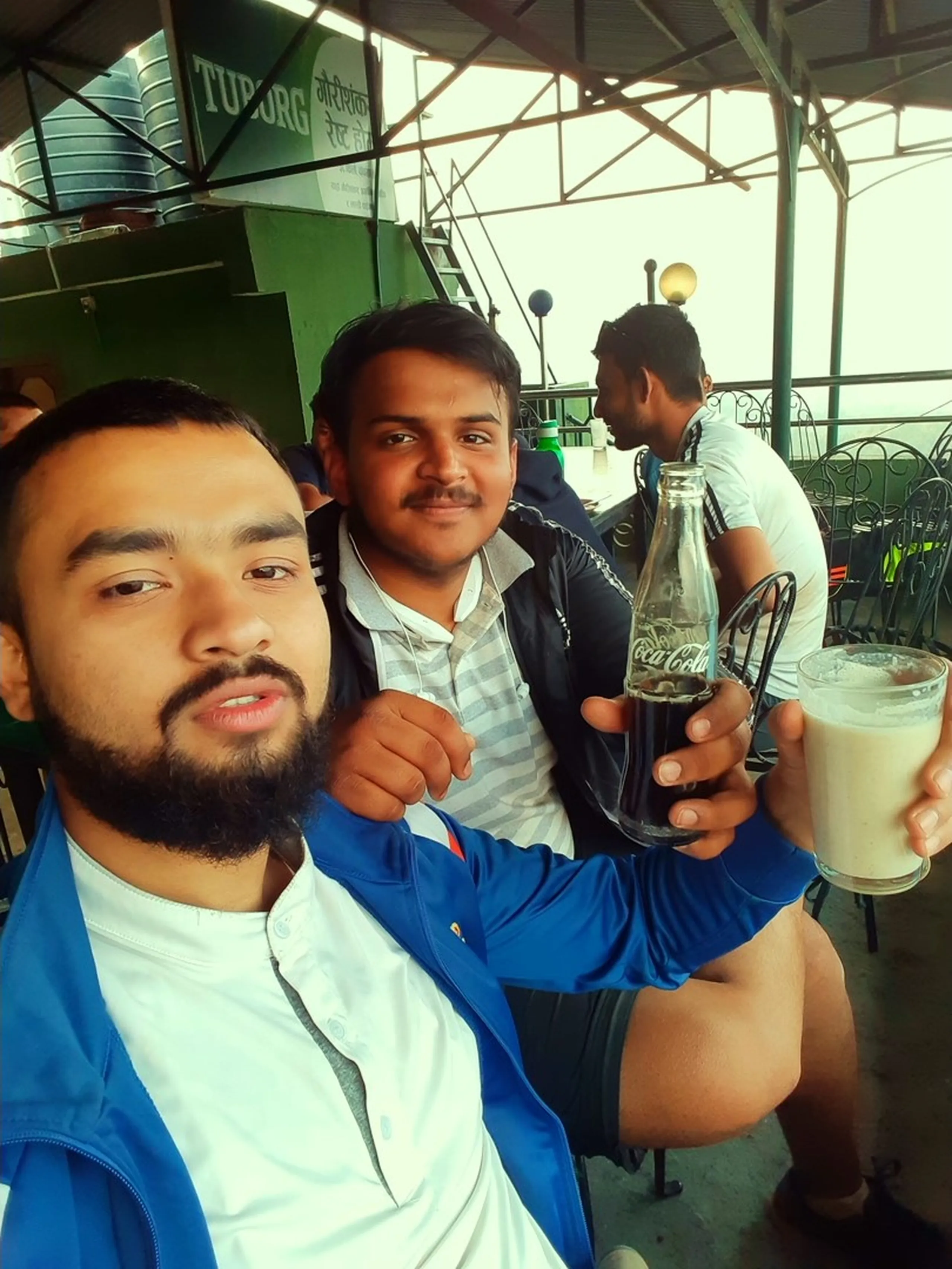 A selfie of two young men sitting at an outdoor rooftop or terrace bar or café. The man in the foreground has a beard and wears a blue jacket; he is taking the photo. The man beside him holds up a glass Coca-Cola bottle in one hand and a large glass of what appears to be a creamy white drink (possibly lassi or a milkshake) in the other, grinning. Behind them, green café signage (possibly a Tuborg beer advertisement), metal awning struts, a lamp, and another person seated in the background are visible. The setting feels like a popular hilltop hangout spot.