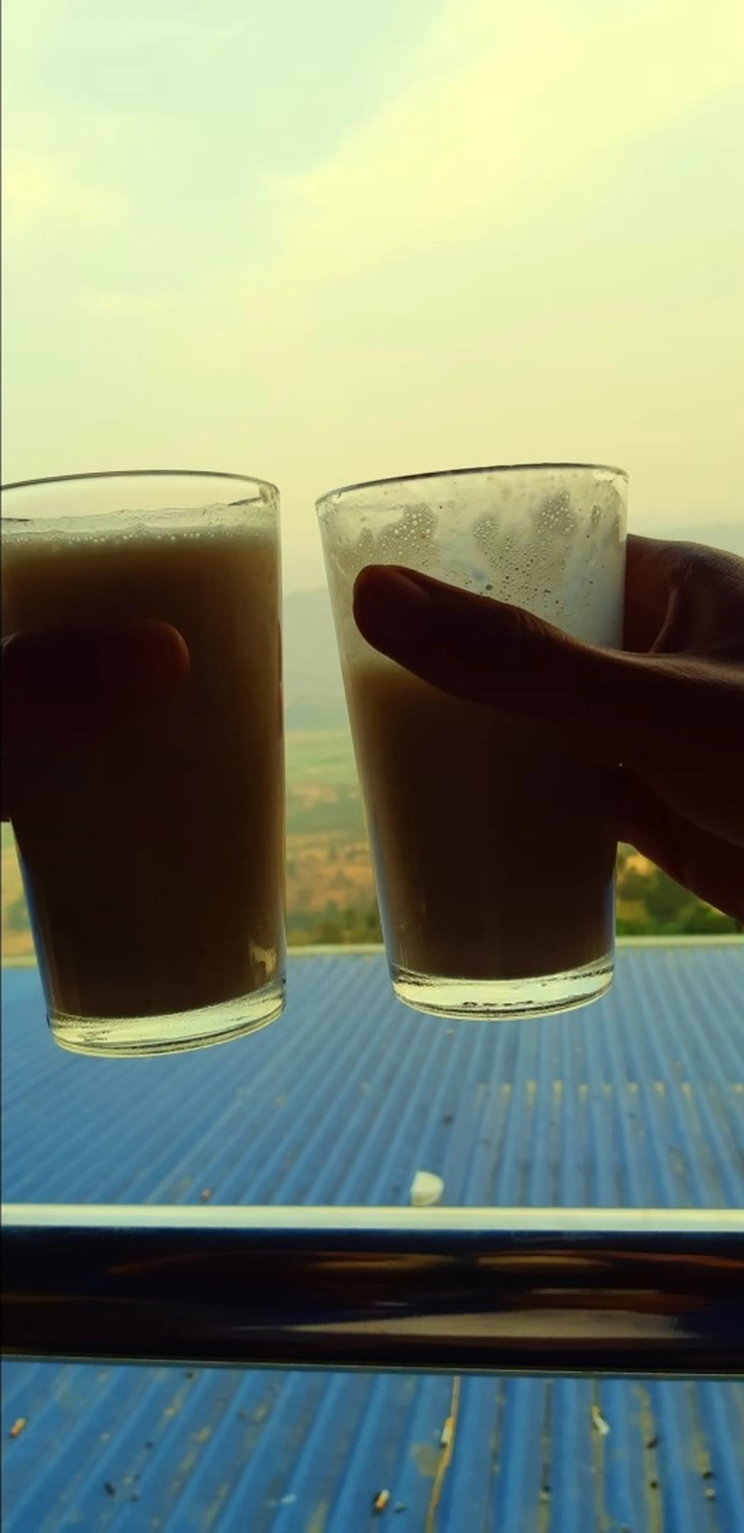 A close-up shot of two hands each holding a glass of milky brown tea (chai), raised toward each other as if clinking in a toast or cheers gesture. The glasses are standard clear cylindrical drinking glasses, slightly fogged and warm-looking. The background reveals a sweeping open landscape - flat green fields stretching into the distance under a pale, hazy yellow-white sky - visible just above a bright blue corrugated metal rooftop railing. The silhouetted hands and glowing backlit sky give the image a cinematic, golden-hour warmth.
