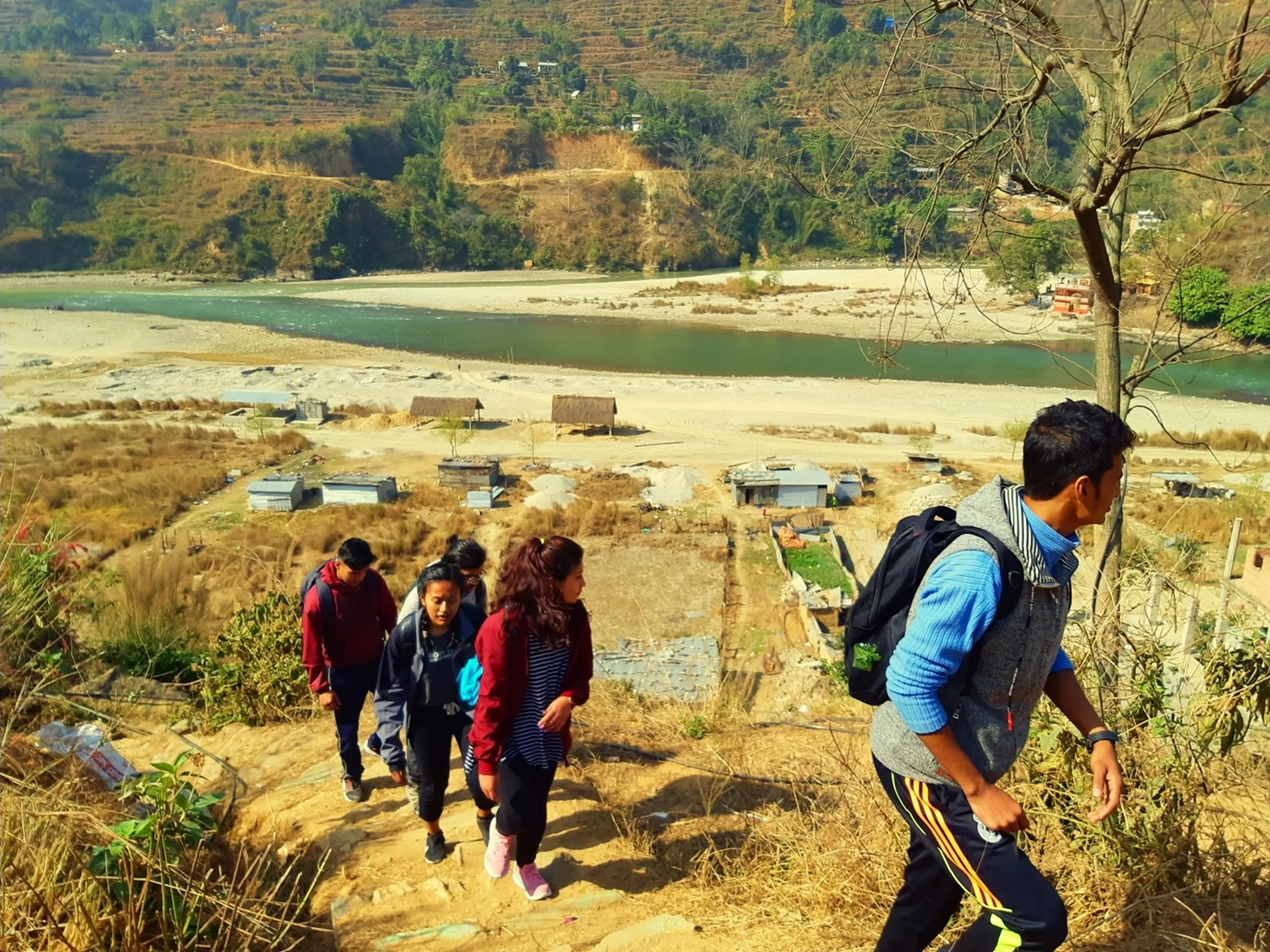 A group of five or six trekkers climb up a dry, scrubby hillside trail, shot from slightly above and ahead of them. The figures are mid-climb, some slightly breathless-looking, all moving upward. Their clothing is colorful - a red jacket, a blue jacket, casual everyday clothes. A bare, leafless tree stands in the right foreground. Below and behind the group, the hillside drops away dramatically to reveal a wide river valley - a broad, pale sandy riverbed with a river running through it, flanked by low sandy banks. Small structures and simple dwellings dot the valley floor beside the river. The view gives the image its scale and drama - the climbers are high above a populated, working river valley.