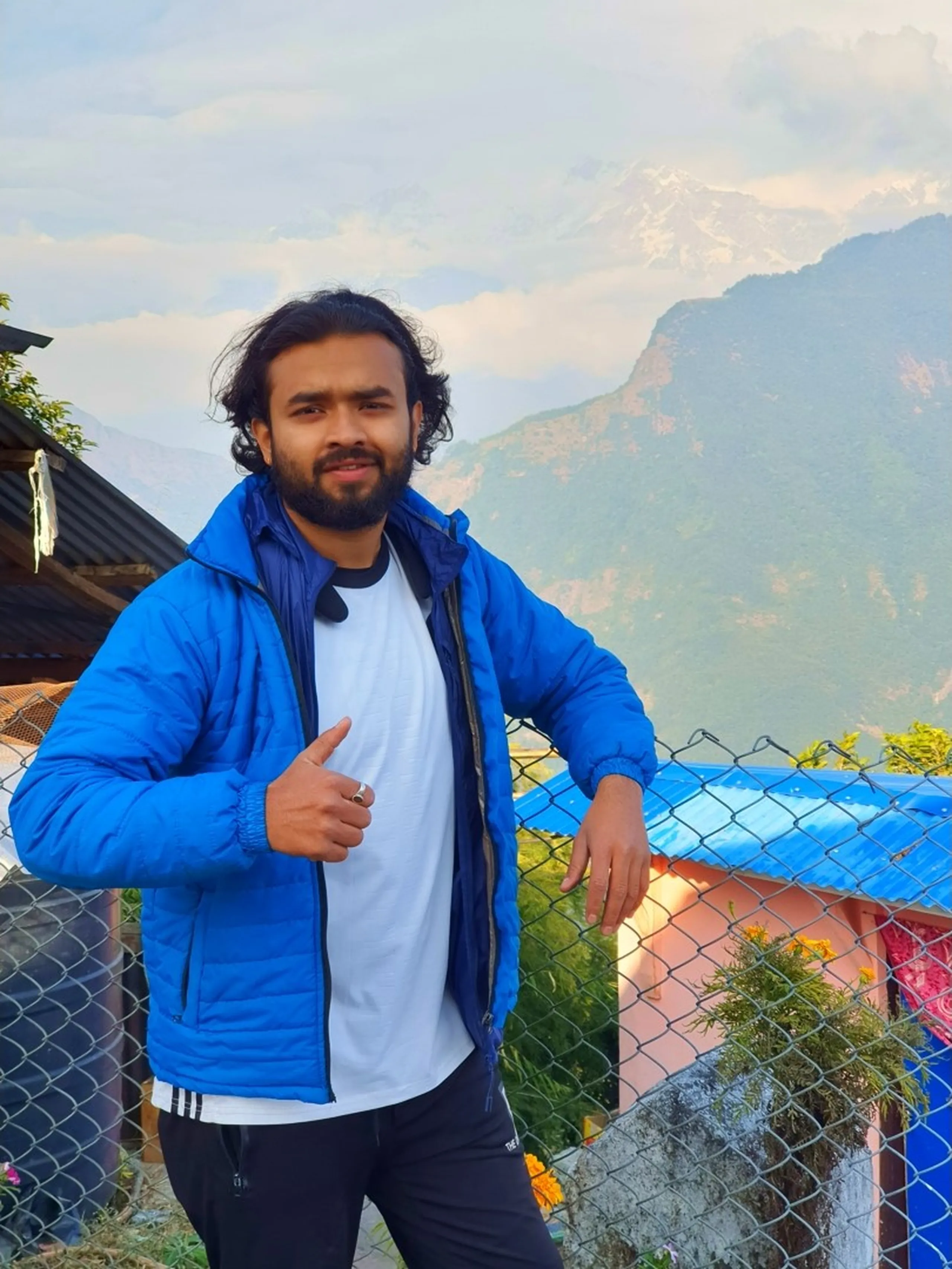 A young South Asian man in his late twenties with a beard, dressed in a vibrant royal blue quilted puffer jacket over a white t-shirt and black track pants, poses confidently with a thumbs-up gesture while leaning one arm against a metal chain-link fence at a mountain viewpoint in Ghale Gaun, Lamjung District, Nepal. Behind him, the spectacular snow-dusted ridgeline of the Annapurna mountain range rises above green forested slopes under a soft cloud-dappled blue sky, with the late afternoon golden light illuminating the scene. Small yellow marigold flowers bloom near the fence base. The photo radiates the joy and accomplishment of reaching a Himalayan trekking destination. Keywords: thumbs up Nepal mountain trek, Annapurna range viewpoint Ghale Gaun, Nepal travel portrait December 2019, Gurung village Lamjung Nepal, trekker celebrating Himalaya, blue puffer jacket Nepal mountain, Annapurna Conservation Area village, Nepal adventure travel photography, Ghale Gaun trek 2019 team.
