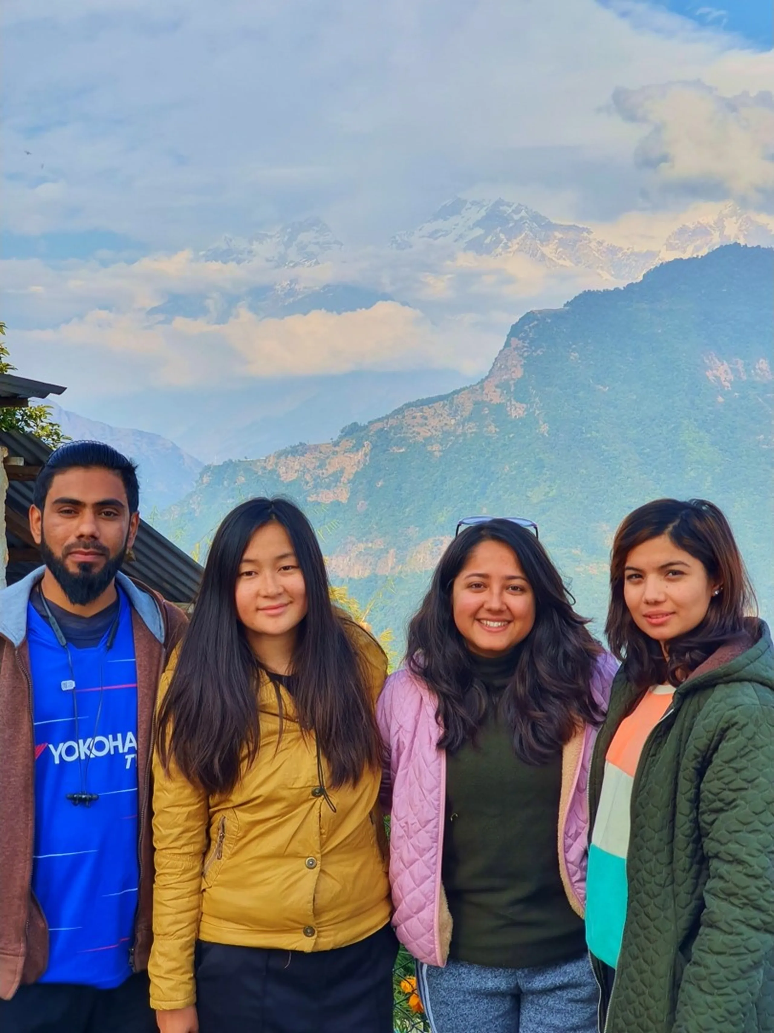 A joyful group portrait of four young trekkers - one man and three women - posing together at a mountain viewpoint in Ghale Gaun, Gurung Village, Lamjung District, Nepal, against a breathtaking backdrop of the snow-capped Annapurna Himalayan range partially veiled in clouds above layered green and rocky mountain ridges under a warm blue sky. The man on the left wears a blue Yokohama-branded jersey with a brown cardigan; the first woman beside him wears a mustard yellow quilted vest with dark pants and has straight long dark hair; the woman in the center wears a black turtleneck under a dusty pink puffer jacket and has wavy dark hair with sunglasses perched on her head; the woman on the right has long dark hair and wears a green and orange striped quilted jacket. All are smiling broadly, capturing the warmth of team camaraderie during a group trek. Keywords: Ghale Gaun group trek Nepal, Annapurna range background group photo, Gurung village Lamjung Nepal team trek, Nepal trekking friends photography, Annapurna Conservation Area group portrait, Nepal corporate team building trek 2019, snow mountain background group photo Nepal, women trekking Nepal Himalaya, Deerwalk team trek Ghale Gaun 2019.