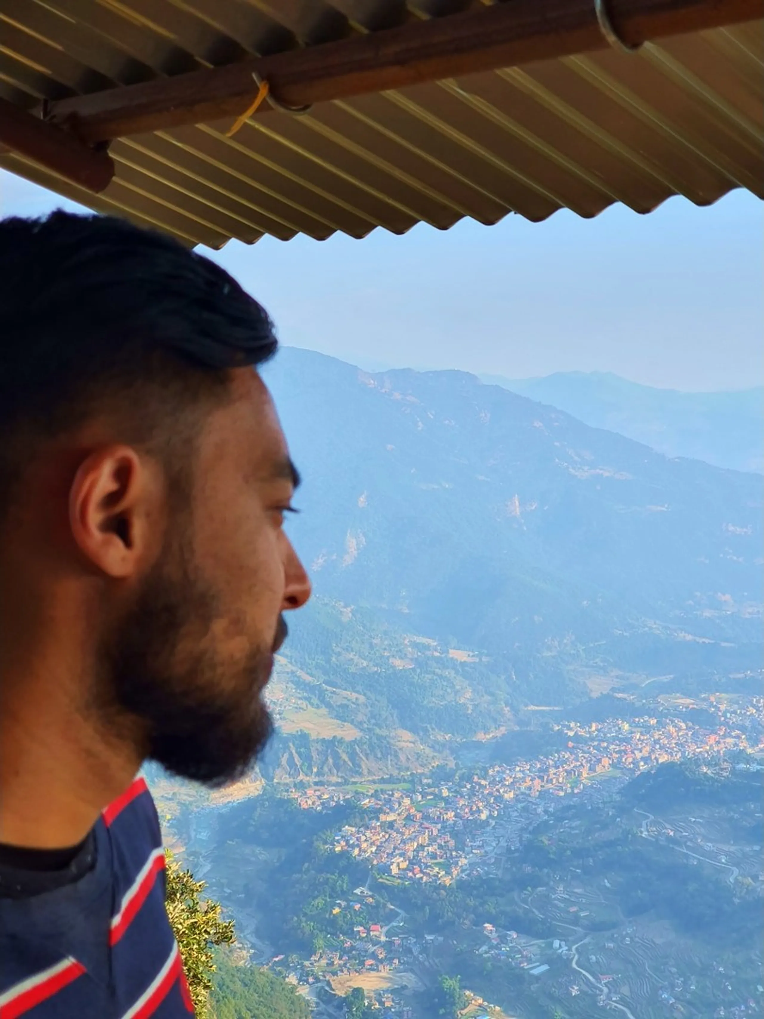 A close-up profile portrait of a bearded South Asian man shot from over his left shoulder as he gazes pensively toward a sweeping aerial panoramic view of Besisahar town, the district headquarters of Lamjung, Nepal, seen far below nestled in the green Marsyangdi river valley between forested mountain ridges fading into a hazy blue morning atmosphere. The man wears a navy blue and red striped jersey, and the low corrugated metal roof eave of a mountain teahouse or lodge frames the top of the image, suggesting he is seated at a high-altitude rest stop on the Ghale Gaun trek. The juxtaposition of the close human figure and the distant town far below vividly conveys the altitude gain of the trek. Keywords: Besisahar view from Ghale Gaun Nepal, Lamjung valley aerial view trekking, Marsyangdi valley Nepal viewpoint, Annapurna region altitude view, Nepal mountain teahouse view, Ghale Gaun elevated viewpoint, Lamjung District panorama Nepal, trekking Nepal altitude photography, bird's eye view Besisahar Nepal 2019.