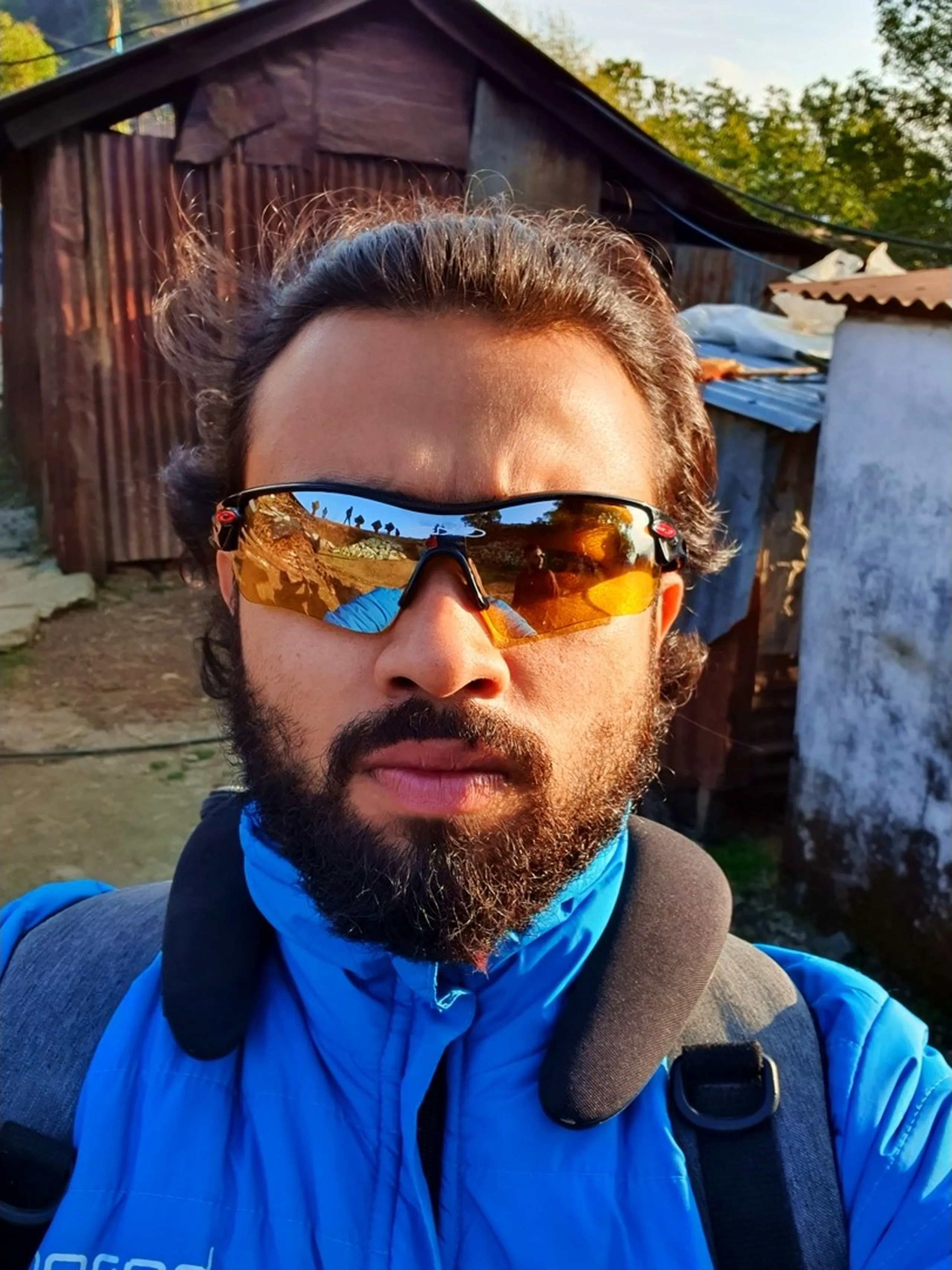 An intense close-up selfie portrait of a bearded South Asian man wearing large wraparound sports sunglasses with striking orange-tinted amber mirrored lenses that reflect the mountain trekking landscape behind him - visible in the reflection are golden dry slopes, blue tarpaulin, and the backs of other trekkers. He has a thick dark beard and curly black hair and wears a blue puffer jacket with a dark grey collar and a trekking backpack strap visible on his right shoulder. The background shows a rustic corrugated iron-roofed village structure amid trees in warm golden morning sunlight, consistent with a mountain village setting in Ghale Gaun, Lamjung District, Nepal. His serious, focused expression embodies the rugged spirit of Himalayan trekking. Keywords: trekking sunglasses selfie Nepal, orange mirror lens sports glasses Nepal trek, Ghale Gaun Gurung village portrait, Nepal trekking gear close-up, Himalayan trek selfie 2019, bearded trekker Nepal mountain village, adventure trekker portrait Nepal, Lamjung District trek photography, Annapurna Conservation Area Nepal.