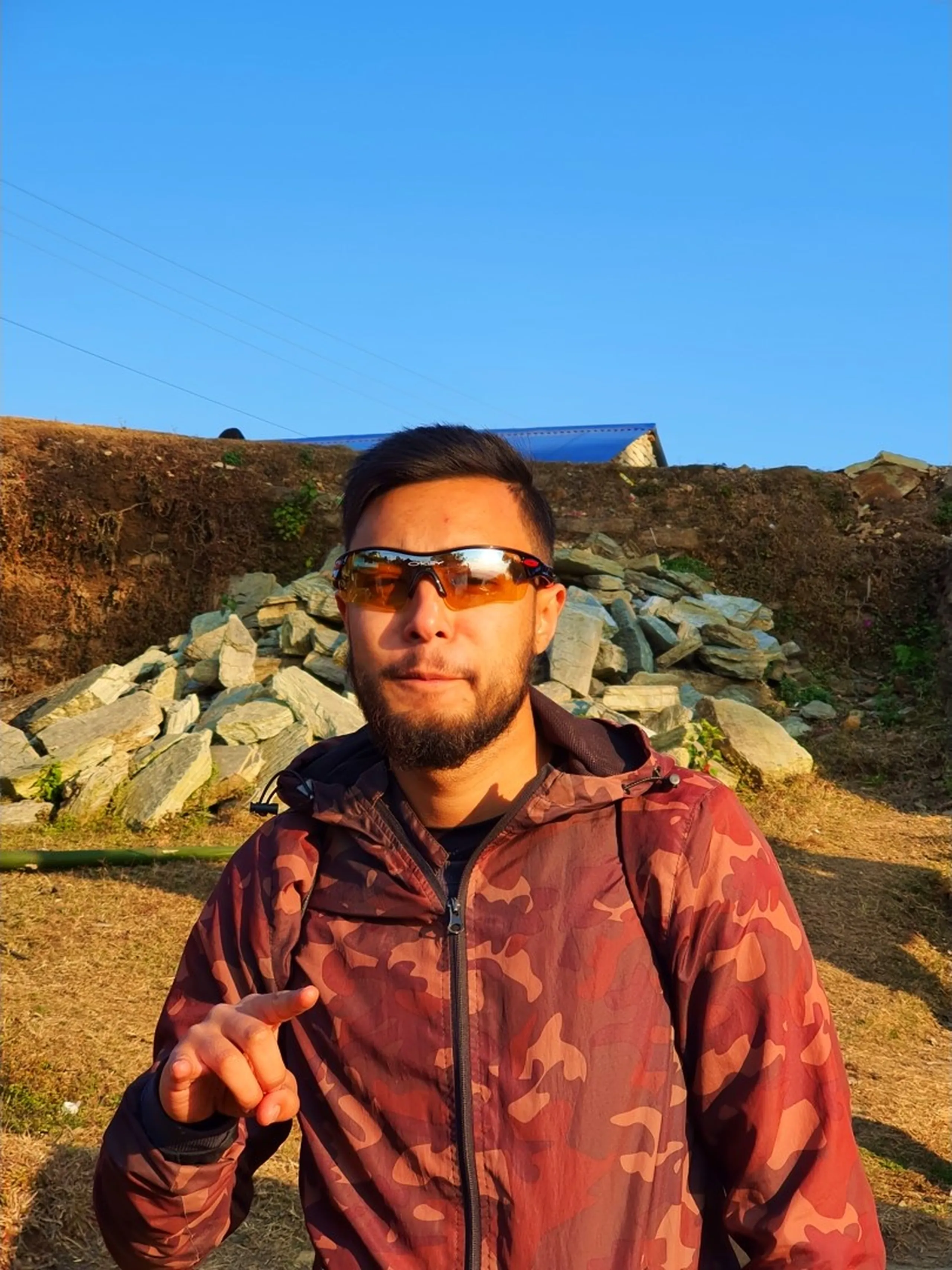 A young East Asian-featured man in his late twenties stands outdoors on dry golden mountain grassland in a high-altitude Himalayan setting near Ghale Gaun, Lamjung District, Nepal, under a flawless cloudless deep blue sky. He wears a brown and khaki camouflage zip-up trekking jacket and sports the same model of orange-tinted wraparound sports sunglasses seen on another group member, suggesting the glasses are being shared among the trekking team. He points one finger directly toward the camera in a playful gesture. Behind him, a low stone wall construction, dry earth, and a small blue-roofed building are visible. The vibrant blue sky and warm golden light give the image a crisp, high-altitude morning feel. Keywords: Ghale Gaun Nepal mountain portrait, high altitude blue sky Nepal trek, camo jacket trekking Nepal, orange sunglasses trekker Nepal, Lamjung Gurung village Nepal, Annapurna Conservation Area trekking, Nepal Himalayan sunshine portrait, group trek Nepal December 2019, trekker pointing camera Nepal.