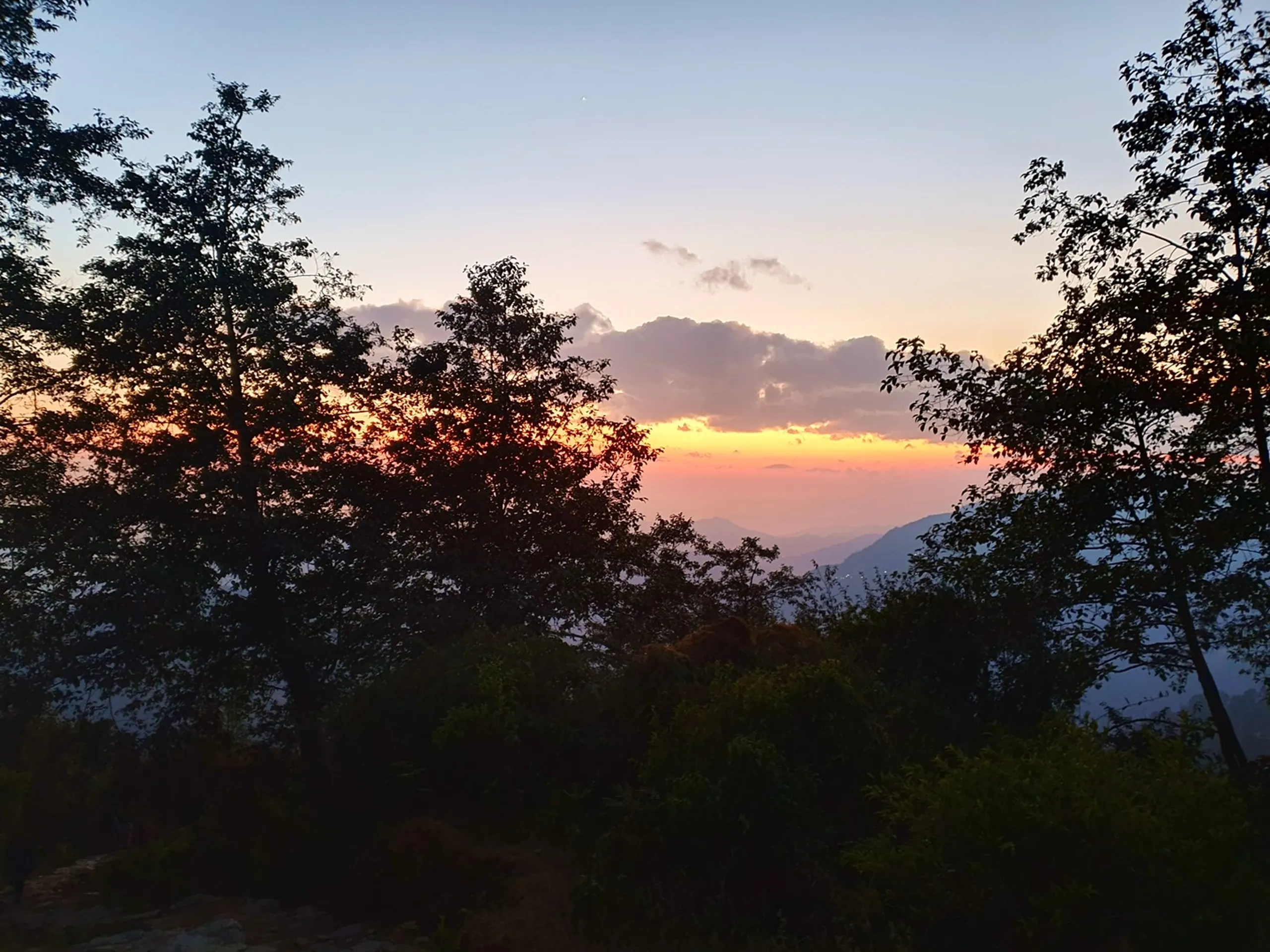 A tranquil and painterly twilight landscape photograph taken from a hillside forest clearing near Ghale Gaun, Lamjung District, Nepal, looking out over successive silhouetted ridgelines of forested mountains fading into a soft lavender and lilac haze in the far distance. The sky is a gradient from pale blue-grey at the top through warm amber and dusty rose near the horizon where the last embers of the setting sun glow behind a central cloud bank tinted pink and gold. Dark tree silhouettes with leafy canopies frame the left and right edges of the frame, creating a natural window effect. A tiny star or planet is faintly visible in the upper sky, and thin mountain lights sparkle in the valley below. Keywords: Ghale Gaun twilight landscape, Nepal mountain sunset silhouette trees, Lamjung District evening photography, Himalayan twilight nature Nepal, Annapurna foothills dusk, mountain silhouette Nepal trek, Nepal travel landscape photography, golden hour Nepal mountains 2019, peaceful Nepal forest sunset, Gurung village Lamjung evening view.