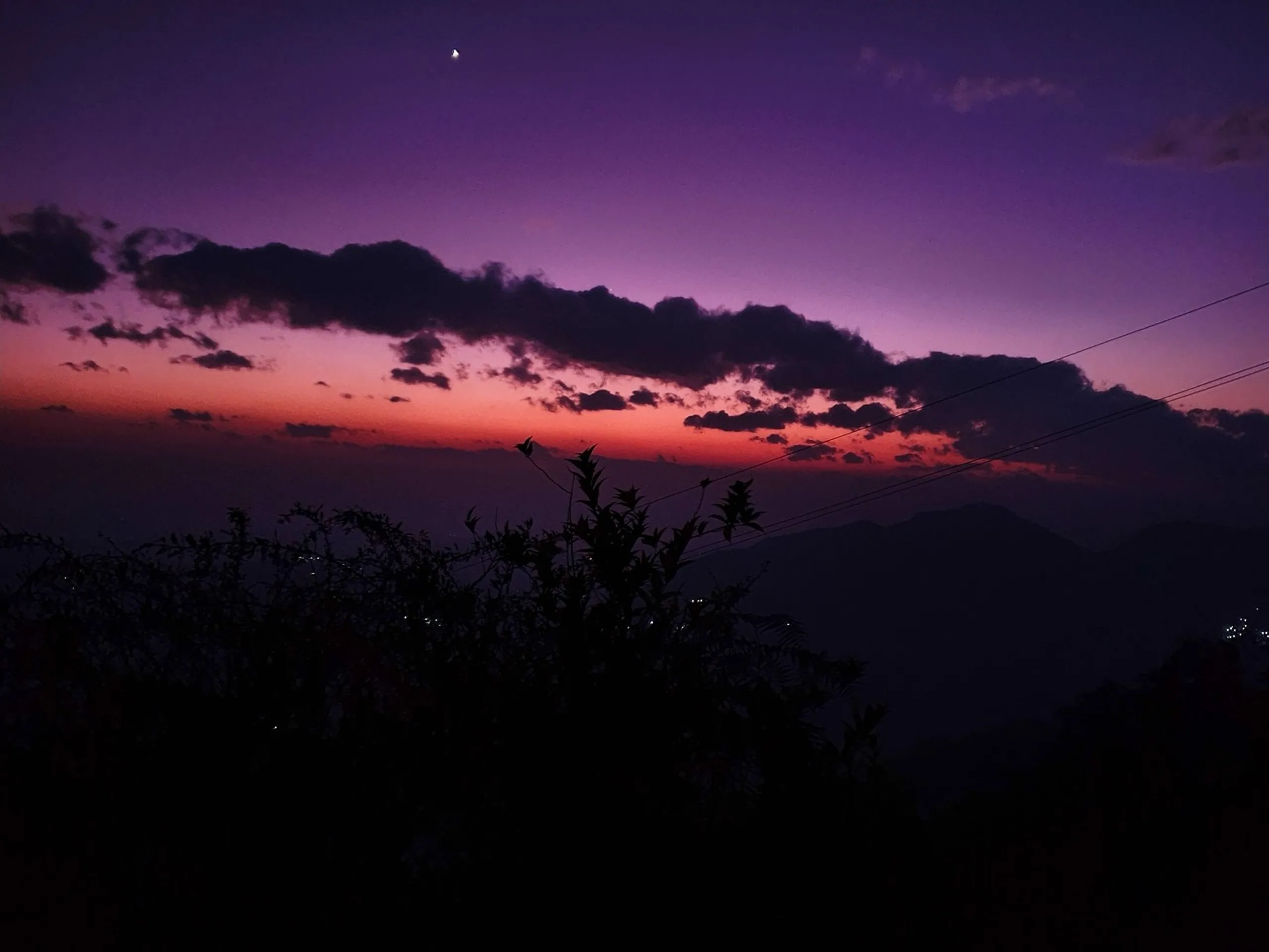 An extraordinarily vivid and cinematic after-sunset sky photograph taken from a hilltop in Ghale Gaun, Lamjung District, Nepal - one of the most striking images of the trek. The sky has transformed into a deep, saturated violet-purple twilight with a crescent moon glowing near the top center. A dramatic horizontal band of turbulent dark clouds stretches across the mid-sky, backlit by an intense fuchsia and crimson afterglow along the horizon that creates a vivid contrast between the dark silhouetted Himalayan ridges below and the luminous purple heavens above. Power line cables cross the upper right corner, and dark silhouetted shrubs and vegetation occupy the lower foreground, their outlines visible against the glowing sky. Distant valley lights twinkle at the base of the mountain silhouettes. Keywords: Nepal purple twilight sky photography, Ghale Gaun after sunset sky, Himalayan twilight purple sky Nepal, crescent moon Nepal mountain night, Lamjung District Nepal night sky, dramatic Nepal sky travel photography, Annapurna region twilight 2019, Nepal nature photography purple, dark clouds glowing horizon Nepal, mountain night sky Nepal trek.