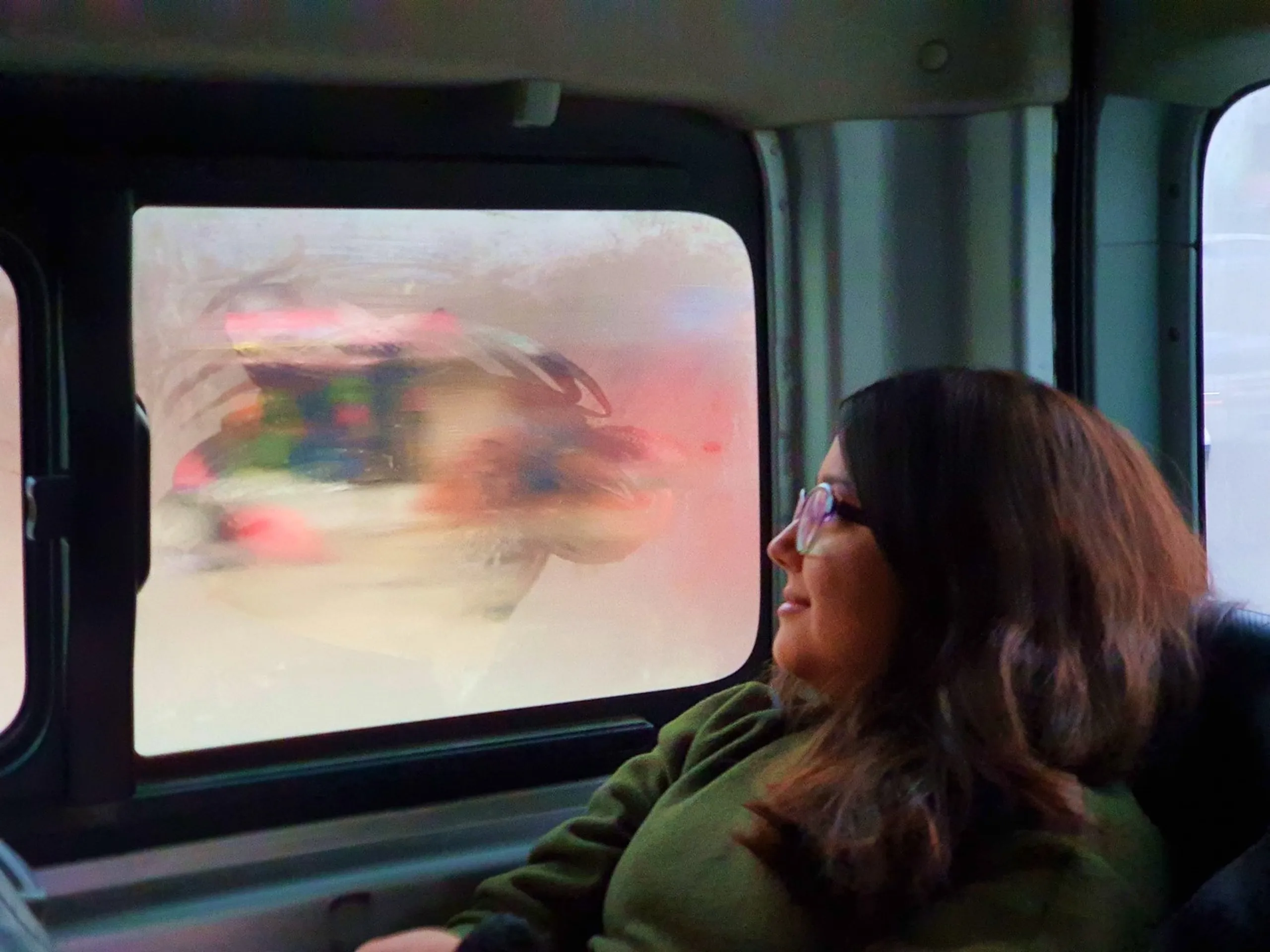 A young South Asian woman with long dark brown hair, wearing glasses with translucent pink frames and a cozy olive green hoodie, sits thoughtfully inside a public bus or microvan in Nepal, gazing out a large fogged-up window. The condensation-covered window glass reflects blurred streaks of colorful traffic lights - red, pink, blue, and white - suggesting movement through a busy Nepali street at dusk or night. The interior of the vehicle is teal-grey, with another window visible on the right. The scene captures the quiet, reflective mood of a team member on their way from Kathmandu toward Ghale Gaun, Lamjung - a poetic moment of wanderlust at the start of a Himalayan trek. Keywords: Nepal bus journey travel photography, Kathmandu to Lamjung road trip, foggy bus window reflections Nepal, woman traveler Nepal trek, Ghale Gaun trek 2019 journey, trekking Nepal December, Annapurna region travel, Nepali public transport night travel.