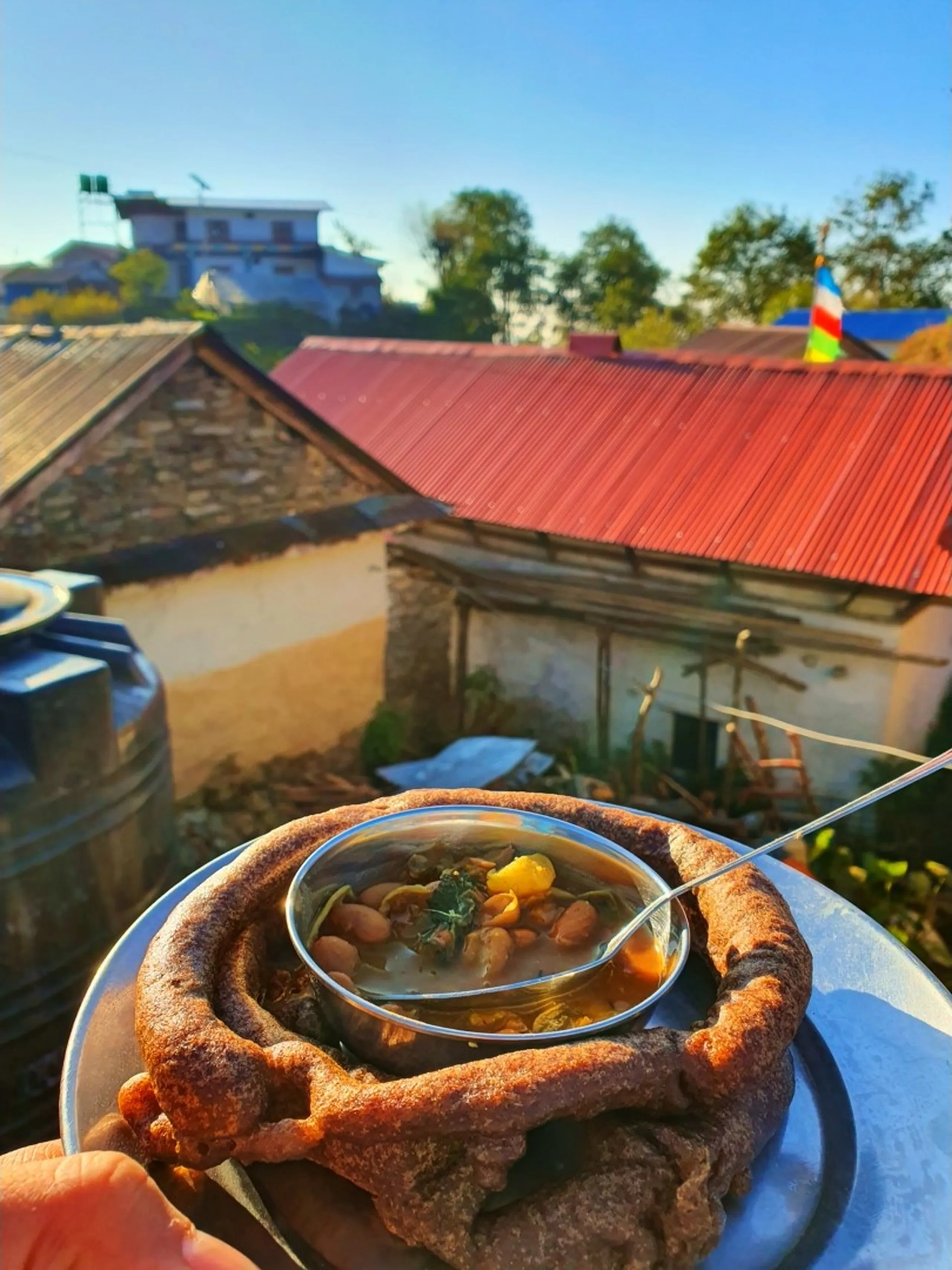 A close-up first-person perspective of a hand holding a round stainless steel plate containing a large, thick, dark-brown ring-shaped deep-fried bread known as 'ghare roti' or traditional Nepali sel roti, with a smaller stainless steel bowl of bean and vegetable soup - likely black-eyed pea curry with leafy greens and potato chunks in a golden broth with a spoon - placed inside the hollow center of the bread. The food is being held up against an outdoor backdrop of traditional Nepali mountain village architecture: a stone-walled building with a rusty red corrugated iron roof, a black plastic water storage tank on the left, and a colorful Buddhist or Hindu triangular prayer flag in the far background near a tree and a white building. The warm golden morning sunlight illuminates the scene, suggesting a fresh mountain breakfast at a homestay in Ghale Gaun, Lamjung District, Nepal. Keywords: traditional Nepali breakfast Ghale Gaun, sel roti bean soup Nepal mountain, Gurung village food Nepal, Nepali mountain homestay breakfast, Nepal local food trekking, ghare roti Nepal recipe, Annapurna Conservation Area food, Nepal trek morning meal, authentic Nepali cuisine Lamjung, Nepal food travel blog 2019.