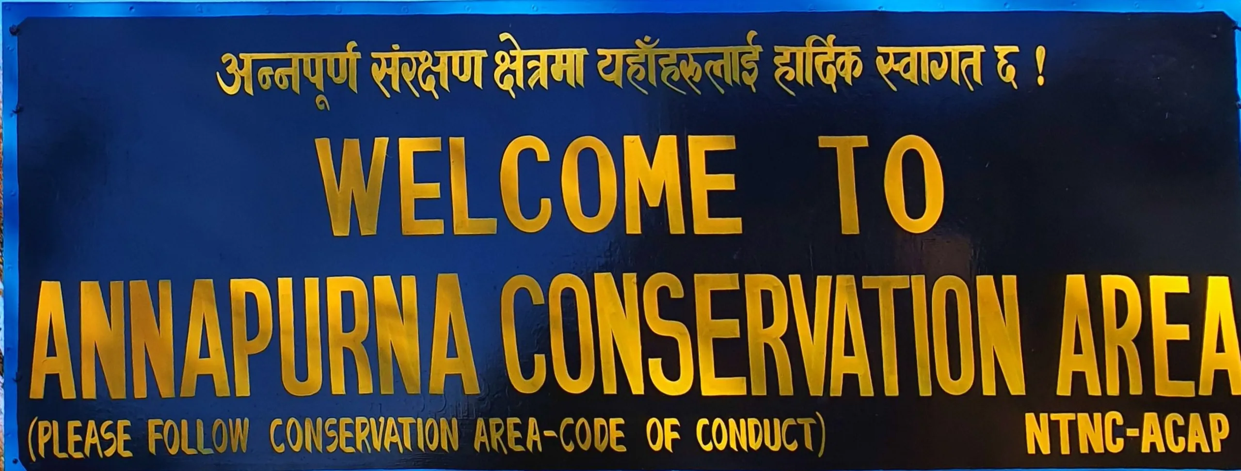 A close-up photograph of a large official dark navy blue signboard with bold bright gold lettering reading 'WELCOME TO ANNAPURNA CONSERVATION AREA' in English, with a secondary line in smaller gold text reading '(PLEASE FOLLOW CONSERVATION AREA-CODE OF CONDUCT)' and the official emblem abbreviation 'NTNC-ACAP' in the bottom right corner. Above the English text, a line of Devanagari script in golden yellow reads the Nepali language welcome message. The sign has a vivid blue border and is photographed straight-on in bright daylight. This is the official entry gateway signboard managed by the National Trust for Nature Conservation under the Annapurna Conservation Area Project - marking the formal entry point into one of Nepal's most celebrated protected natural areas, through which the Ghale Gaun trek route passes in Lamjung District. Keywords: Annapurna Conservation Area entry sign Nepal, NTNC ACAP signboard Nepal, Annapurna trekking entry point, Nepal conservation area sign, Ghale Gaun trek entry Lamjung, Annapurna region trekking permit, Nepal protected area signboard, ACAP Nepal tourism, Annapurna trekking rules Nepal, Nepal trekking signboard 2019.