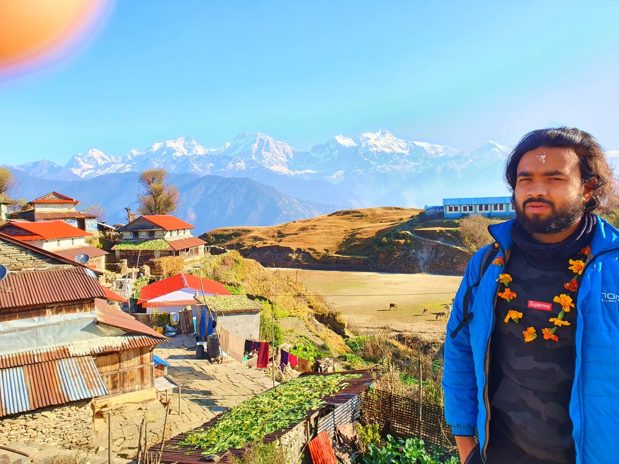 A portrait of the same bearded South Asian man with long dark hair, wearing a blue puffer jacket over a black Supreme sweatshirt and a traditional orange marigold flower garland with a white tika on his forehead, standing confidently in front of the sweeping panorama of Ghale Gaun, the iconic Gurung village in Lamjung District, Nepal. The village spreads across the hillside behind him - a cluster of traditional stone houses with bright red corrugated metal roofs, satellite dish, stone walls, vegetable garden plots, drying laundry, and a blue tented structure. Behind the village, a broad golden-dry grassy plateau slopes upward where two small animals - likely horses or donkeys - graze freely near a blue-roofed building. Above this, the majestic snow-capped ridgeline of the Annapurna Himalayan massif stretches across the entire upper background under a clear blue sky, with a subtle lens flare visible in the upper left corner from the bright morning sun. Keywords: Ghale Gaun panoramic view Nepal, Annapurna Himalaya behind Gurung village, marigold garland tika welcome Nepal, Lamjung District Nepal village portrait, Ghale Gaun red roof stone houses, Annapurna range Lamjung Himal backdrop, Nepal Gurung village culture trek, Annapurna Conservation Area village trek 2019, Nepal mountain village scenery, trekker Ghale Gaun welcome ceremony.