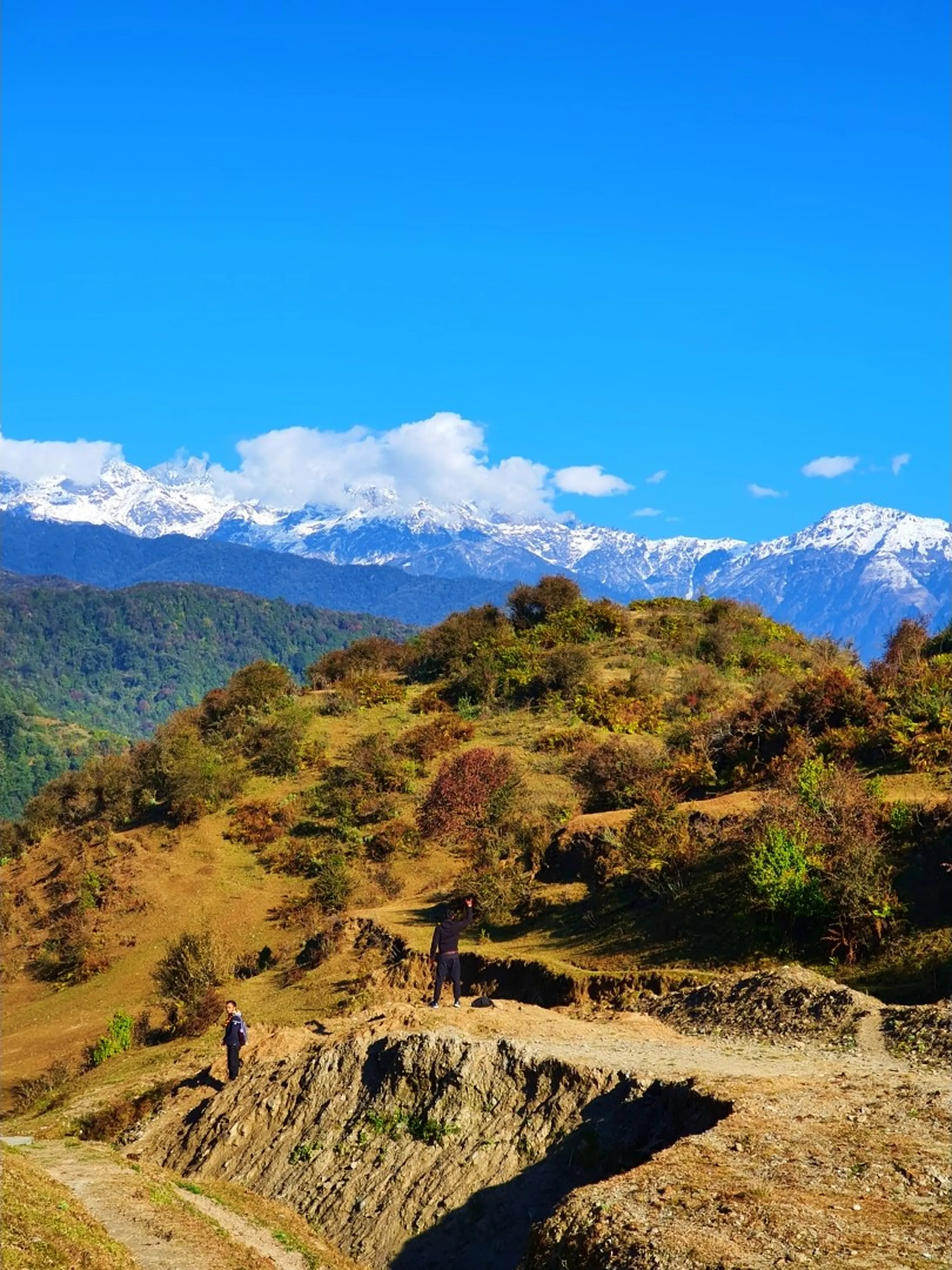 A wide landscape photograph taken on an open hillside trail above Ghale Gaun in Lamjung District, Nepal, showing two tiny human figures - one in dark clothing standing on a rocky ledge in the lower foreground and another in dark clothing further uphill - dwarfed by the vast natural terrain around them. The hillside in the middle ground is covered in dry golden autumn grass, sparse shrubs, and patches of orange and brown seasonal foliage, with a narrow rocky dirt track cutting through the terrain. In the upper half of the image, the spectacular unobstructed panorama of the snow-covered Annapurna Himalayan range stretches across the full width of the frame, its brilliant white glaciated peaks gleaming against a saturated deep blue winter sky dotted with a few white clouds. The scale of the two tiny figures against the massive mountain backdrop dramatically conveys the grandeur of the Himalayan trekking experience. Keywords: Ghale Gaun hillside trail Nepal, Annapurna range panorama trekking, Lamjung District autumn hillside Nepal, two trekkers Himalayan landscape, Nepal mountain trail panorama, Annapurna Conservation Area trail view, snow capped Himalaya Nepal December, tiny figures vast mountain Nepal, Ghale Gaun trekking trail 2019, Annapurna massif full panorama Nepal.