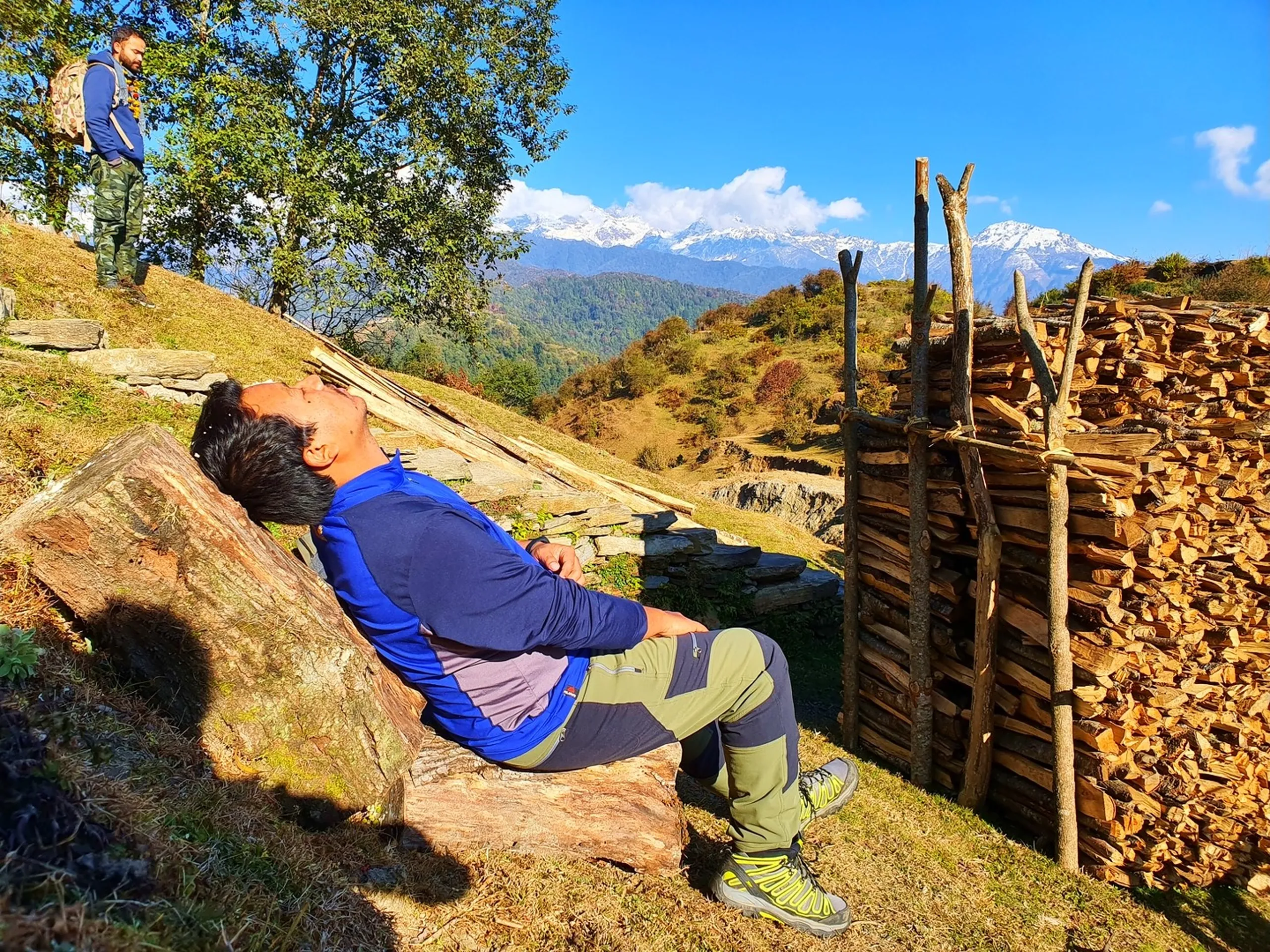 A candid and humorous mid-trek rest moment photographed in warm golden sunlight on a hilltop above Ghale Gaun, Lamjung District, Nepal. In the foreground, a young South Asian man wearing a blue and grey trekking fleece, olive-green trekking pants, and yellow-laced trekking boots reclines completely horizontally against a large rounded brown boulder, his head tilted back and eyes closed as if exhausted or basking in the sunshine - the universal posture of a tired trekker taking a blissful break. A large neat stack of split firewood supported by rustic wooden poles is arranged to his right. In the upper left background, a second group member - wearing a blue jacket with a marigold garland and carrying a camouflage backpack - can be seen standing upright on a higher grassy slope under green trees. The sweeping Annapurna snow peaks and forested Lamjung valleys are visible in the far background. Keywords: trekker resting Nepal mountain, Ghale Gaun trail rest stop, Annapurna trek rest break Nepal, Nepal mountain hiking rest, tired hiker Nepal Himalaya, Lamjung District trekking break, firewood stack Gurung village Nepal, Annapurna Conservation Area trek rest, Nepal winter trekking December 2019, team trek Nepal candid moments.