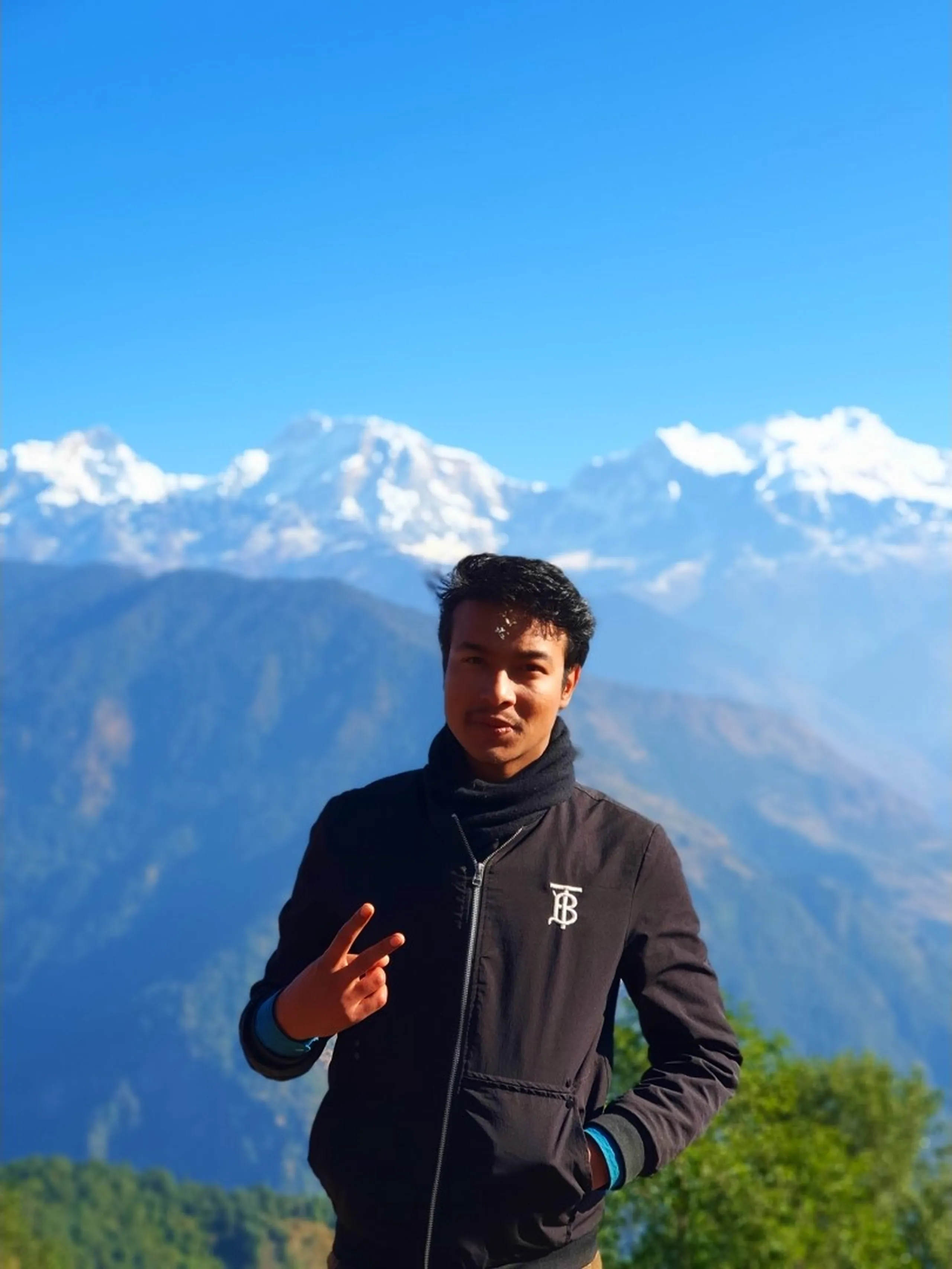 A clean portrait photograph of a young East Asian-featured Nepali man in his late twenties, clean-shaven with short black hair slightly windswept, wearing a smart black zip-up jacket with a blue inner collar and cuffs and a small embroidered monogram logo on the chest, making a cheerful peace sign gesture with his right hand while smiling and looking slightly off-camera. He stands at a high-altitude viewpoint above Ghale Gaun with the breathtaking panoramic backdrop of the snow-covered Annapurna Himalayan peaks - including the broad Annapurna massif and Lamjung Himal - rising dramatically above layered hazy blue-green forested ridges under a cloudless vivid blue sky. A glimpse of green tree leaves is visible in the lower right corner. The crisp mountain air and brilliant scenery behind him perfectly capture the rewarding feeling of reaching the Ghale Gaun trek viewpoint. Keywords: peace sign trekker Nepal mountain, Annapurna range viewpoint portrait Nepal, Ghale Gaun trek viewpoint, Lamjung Himal Annapurna backdrop, Nepal trek portrait clear sky, Gurung village trek Nepal, Nepal mountain travel portrait 2019, Annapurna Conservation Area viewpoint, snow mountain backdrop portrait Nepal, Ghale Gaun hiking success.
