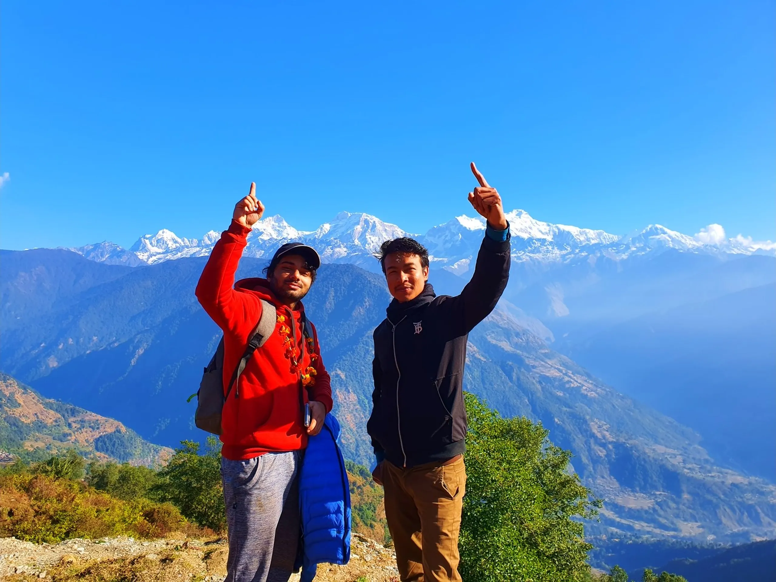 An exuberant and joyful double portrait of two young South Asian men standing side by side at the summit viewpoint above Ghale Gaun, Lamjung District, Nepal, both raising one arm high and pointing a single finger upward toward the sky in a triumphant celebratory gesture. The man on the left wears a bright red hoodie, a dark cap, trekking backpack straps, and a marigold flower garland around his neck, and holds a blue sleeping bag roll in his other hand. The man on the right wears a black zip-up jacket with a blue inner collar and carries nothing, grinning broadly. Behind them, the extraordinary panorama of the Annapurna Himalayan range - with multiple snow-clad peaks including Annapurna II and Lamjung Himal - fills the upper background in brilliant white against a perfectly clear deep blue sky. The lower background shows layers of colorful autumnal forested ridges in shades of gold, green, and brown, and far to the lower right, the faint outline of Besisahar town in the Lamjung valley below can be seen in the haze. Keywords: two trekkers celebrating Nepal mountain, Ghale Gaun summit viewpoint, Annapurna range triumphant pose Nepal, Besisahar valley view from above, Lamjung District viewpoint Nepal trek, Nepal mountain team celebration 2019, pointing sky Annapurna backdrop, Ghale Gaun trek achievement, Nepal trekking friends portrait, Annapurna Conservation Area summit view.