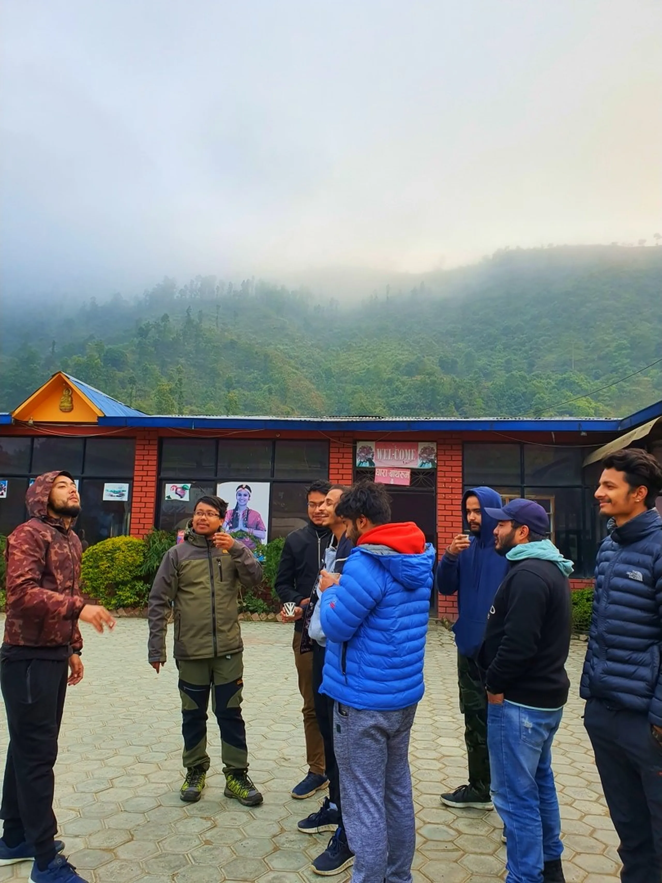 A lively group of eight young Nepali men dressed in trekking jackets and outdoor gear - including blue, red, black, and camo puffer jackets - stand in a semi-circle on a stone-paved courtyard outside a low brick building with a blue corrugated roof and a 'Welcome' sign written in both English and Nepali Devanagari script. A painted portrait of a traditionally dressed Gurung woman is visible on the building's facade, hinting at the indigenous cultural heritage of the region. Mist-covered green forested hills rise dramatically behind the building under an overcast grey sky, signaling the misty mountain atmosphere of Ghale Gaun, Lamjung District, Nepal. The group appears animated, mid-conversation - a snapshot of team bonding before the trek begins. Keywords: Ghale Gaun welcome sign Nepal, Gurung village Lamjung trekking, Nepal group trek 2019, Annapurna Conservation Area trekkers, misty mountain village Nepal, team trekking Nepal Himalaya, Deerwalk trek Ghale Gaun, corporate team trek Nepal, foggy Himalayan village morning.