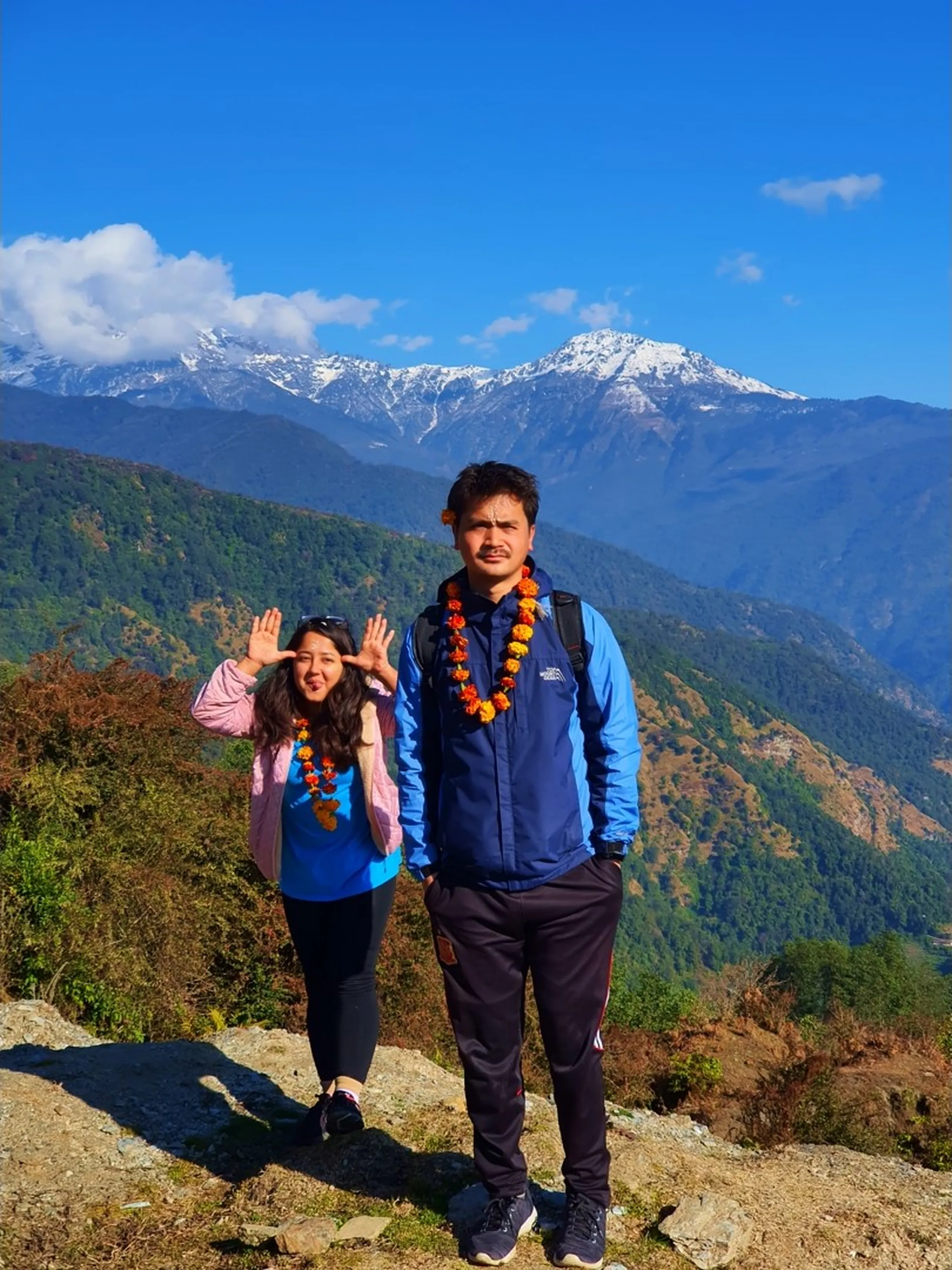 A lively and playful portrait of a young Nepali couple - a woman and a man - posing together at a rocky mountain viewpoint on the Ghale Gaun trek trail in Lamjung District, Nepal. The man stands in the foreground wearing a navy blue two-tone waterproof trekking jacket, dark track pants, and a traditional orange-red marigold flower garland with a trekking backpack - the same man seen earlier in front of the Annapurna snow peaks. The woman behind him wears a light pink puffer jacket, a blue t-shirt, black leggings, and also sports an orange marigold garland; she playfully holds both hands up near her face with fingers spread in a fun surprised expression and sticks out her tongue cheekily while sunglasses rest on top of her head. Behind both of them, layered green and golden-brown forested mountain ridges rise toward a snow-dusted Himalayan peak visible clearly against the blue sky with white clouds. The Besisahar valley in Lamjung fades into the hazy distance on the right. Keywords: Nepal trekking couple marigold garland, Ghale Gaun trek fun photo, Annapurna foothills couple portrait, Lamjung District trek playful, Nepal mountain fun trekking moment, marigold welcome garland Nepal trek, Gurung village trek couple, Besisahar valley backdrop Nepal, Annapurna Conservation Area couple photo, Nepal corporate trek team fun 2019.