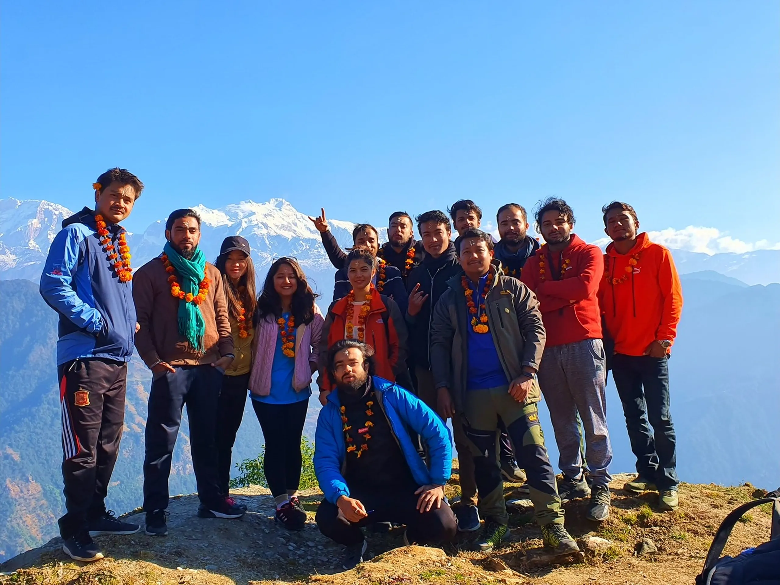 The definitive group photograph of the entire Ghale Gaun trekking team - approximately thirteen young Nepali men and women - gathered together at the high-altitude mountain viewpoint above the Gurung village of Ghale Gaun in Lamjung District, Nepal, with the spectacular snow-capped Annapurna Himalayan range stretching majestically across the full upper background under a vivid blue sky. The group is a vibrant mix of personalities and outfits: many wear traditional orange marigold flower garlands indicating a warm Gurung welcome ceremony. Clothing colors include blue, brown, black, grey, green, orange, and red; some wear trekking boots and backpacks. One person crouches in the center foreground, and others strike fun poses - peace signs, crossed arms, pointing - capturing the euphoric energy of reaching a Himalayan viewpoint together. The valley far below shows layered forested ridges and hazy blue mountains fading into the distance. This is the quintessential Nepal corporate team trek group photo. Keywords: Ghale Gaun group trek photo Nepal, Annapurna range team photo Lamjung, Nepal corporate team building trek, Gurung village marigold garland group, Ghale Gaun summit group portrait 2019, Annapurna Conservation Area team photo, Nepal company trek Deerwalk, snow mountain team photo Nepal, Lamjung District trek group, Nepal Himalayan trek group celebration.